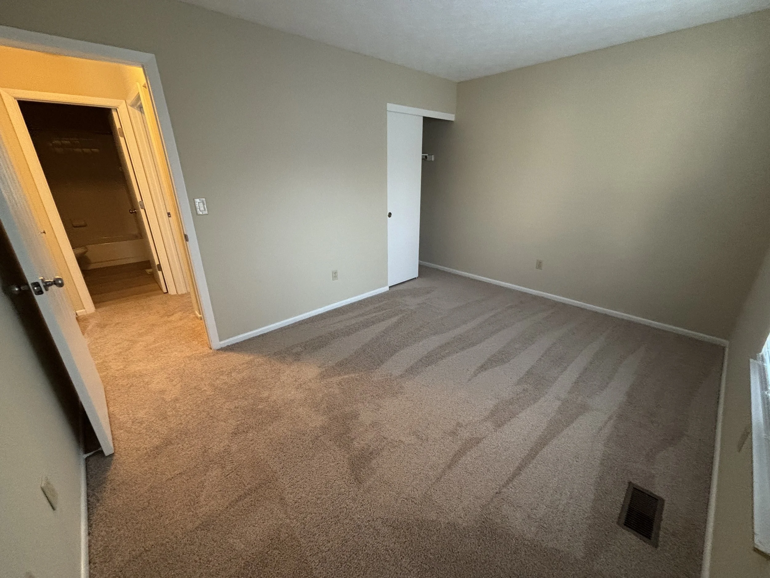 Empty beige carpeted bedroom with a small closet and a window, with a view into the adjacent bathroom.