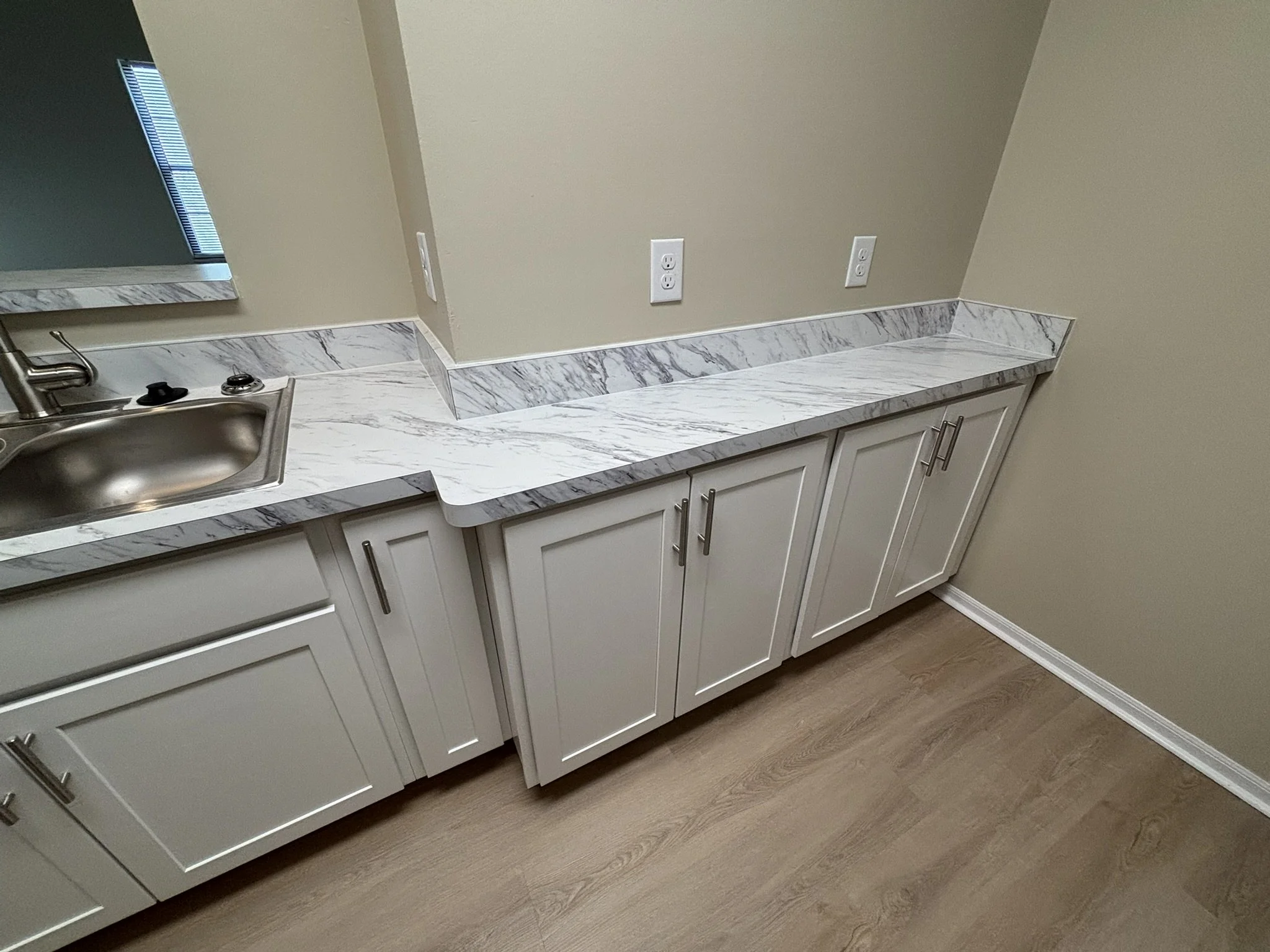 Kitchen counter with marble surface, white cabinetry, a small stainless steel sink, and beige wall with electrical outlets.