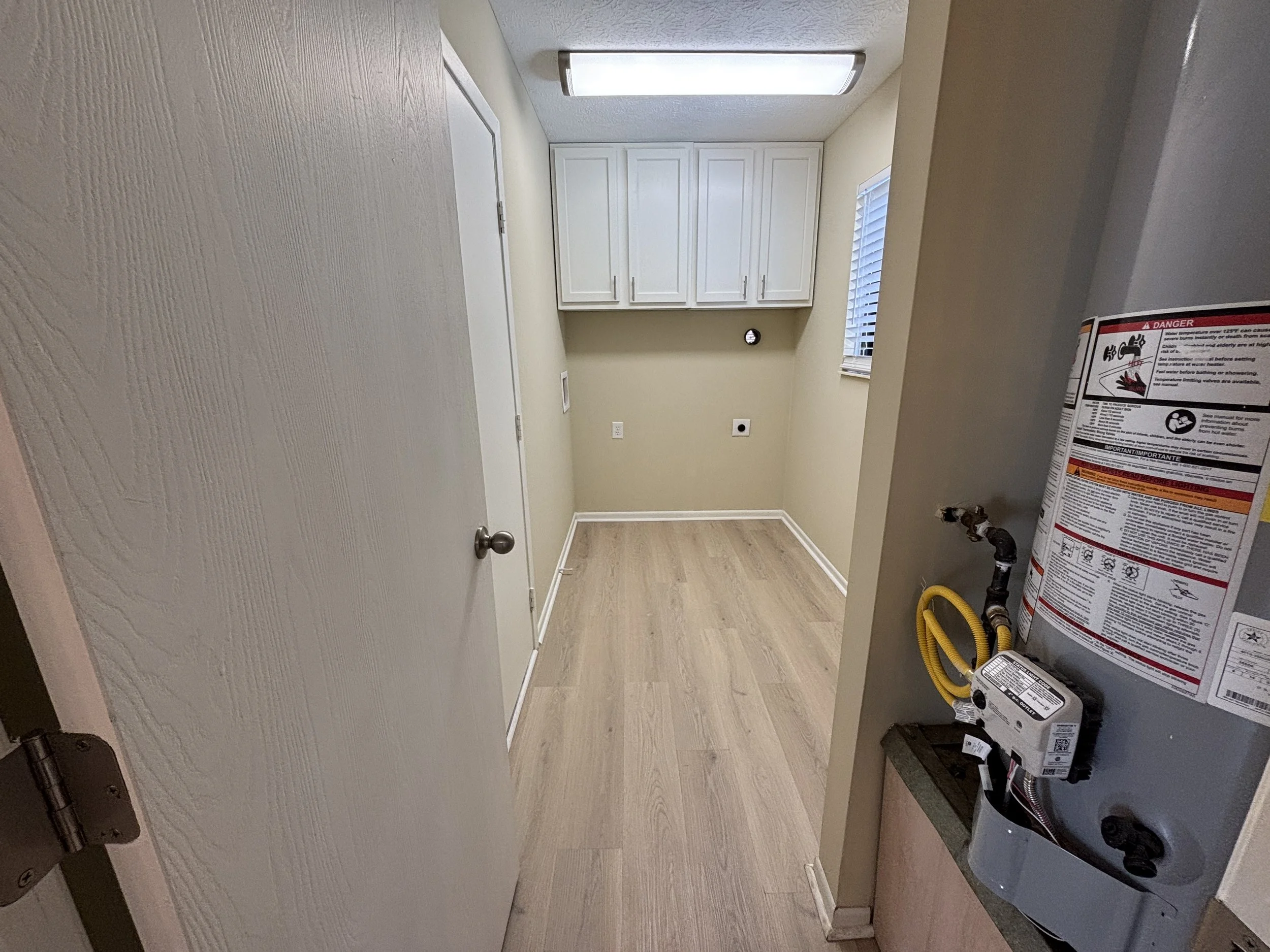 Empty laundry or utility room with white cabinets, beige walls, a window with blinds, and a water heater with yellow hose on the right side.