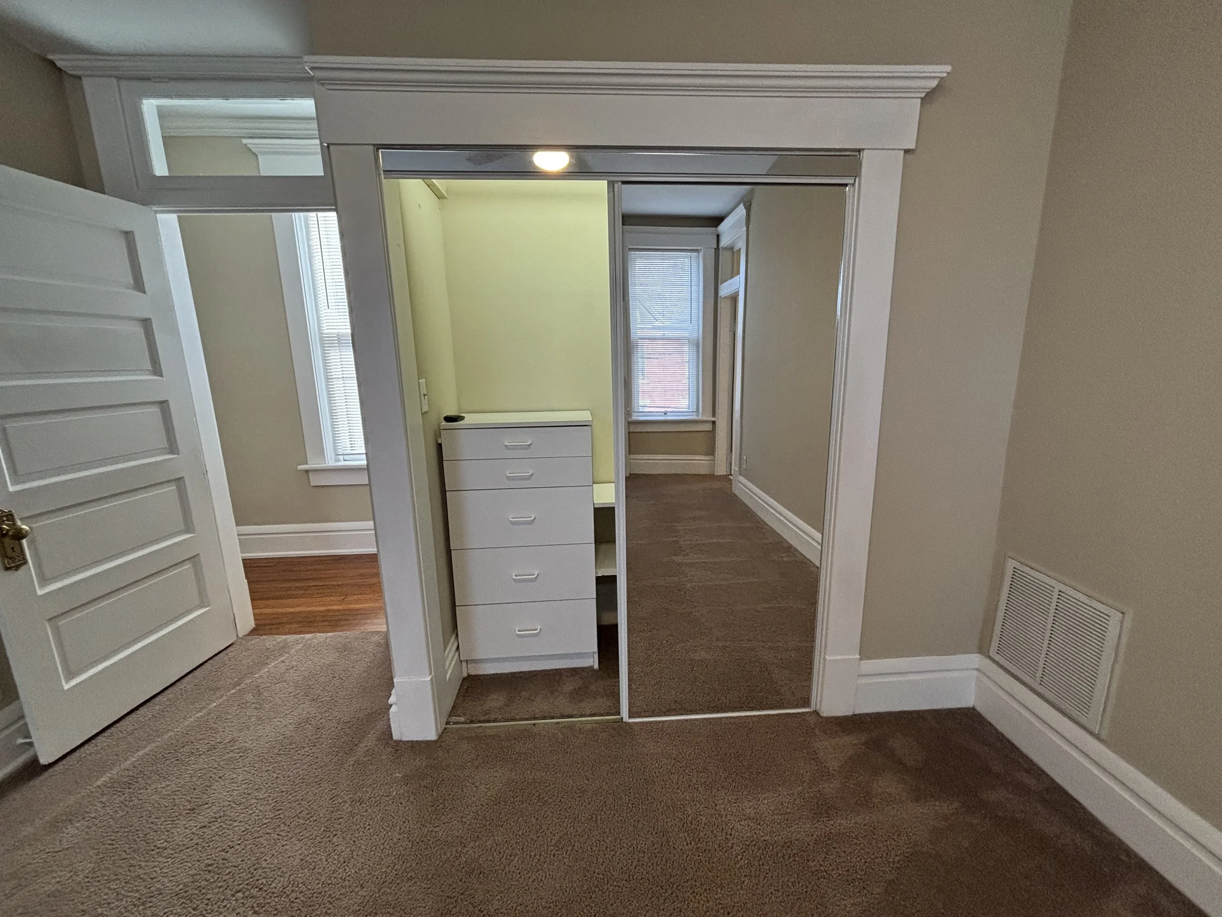 A room with beige walls, a sliding mirror closet door, a small white dresser inside the closet, a window with blinds, and a vent on the wall.