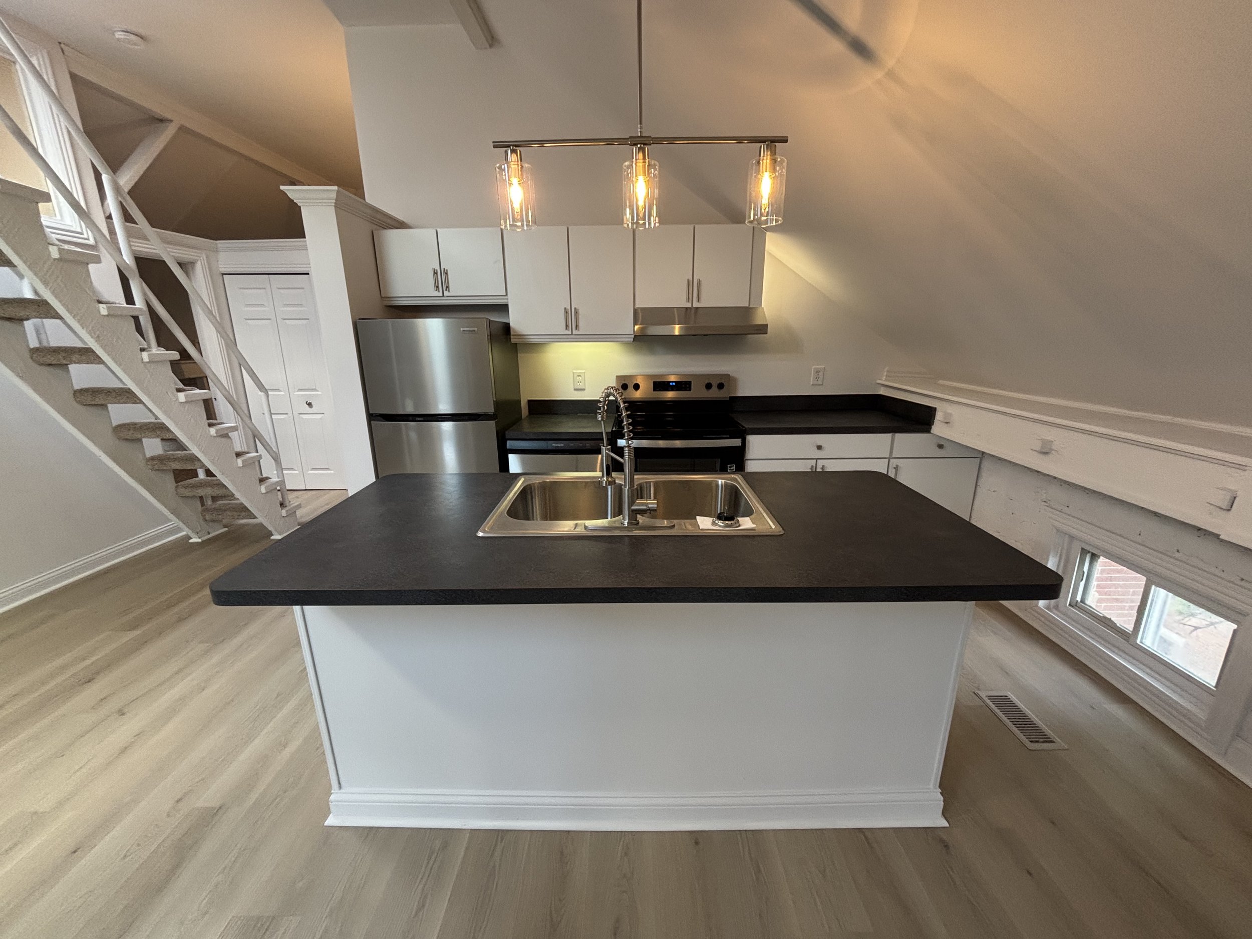 A modern kitchen with white cabinets, stainless steel appliances, and black countertops. There is a kitchen island with a double sink and a gooseneck faucet. A contemporary light fixture hangs above the island, and a window is on the right side of th