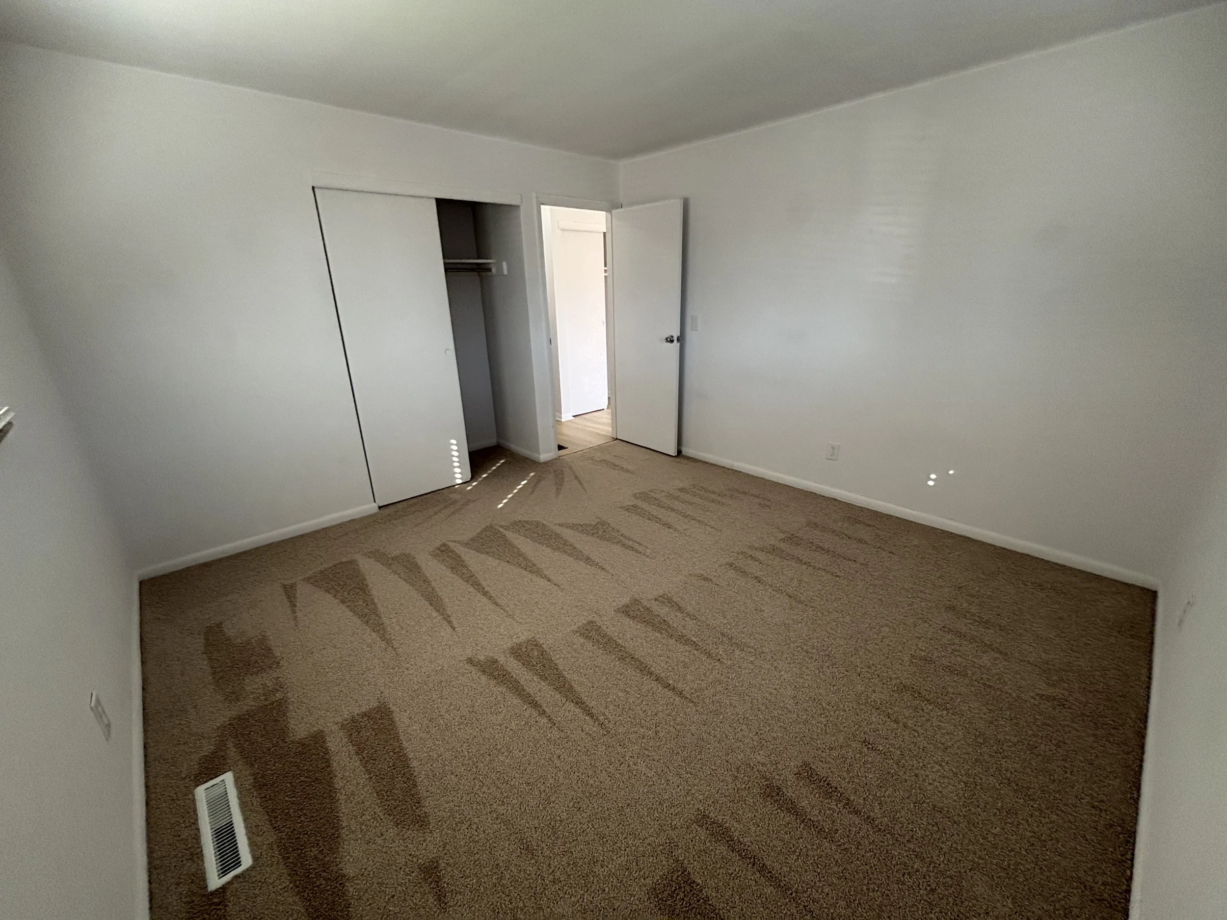 Empty bedroom with beige carpet, white walls, and an open closet door. Sunlight casts patterns on the carpet. There is a door leading to another room or hallway.