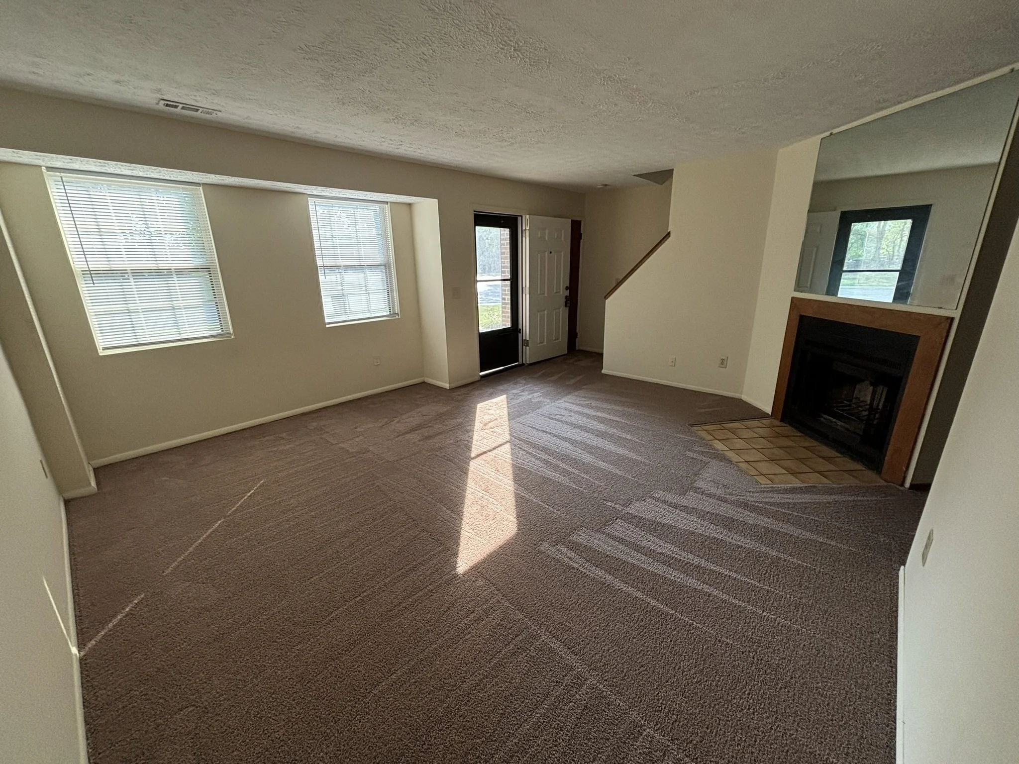 Empty living room with beige carpet, three windows with blinds, a fireplace with a wooden frame, a glass door leading outside, and a staircase.