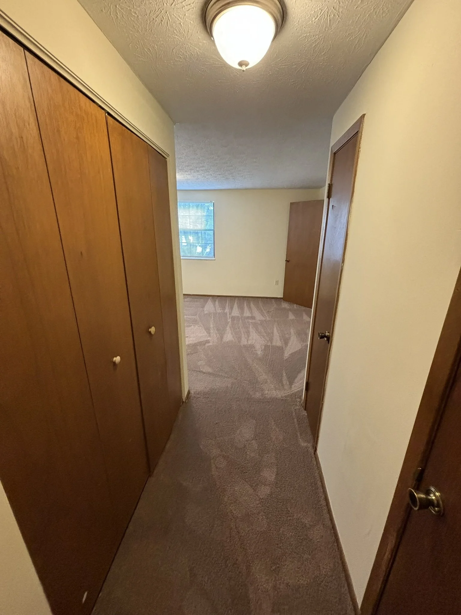 A hallway leading into a bedroom with a window, beige carpeted floor, wooden closet doors on the left, and a closed wooden door on the right.