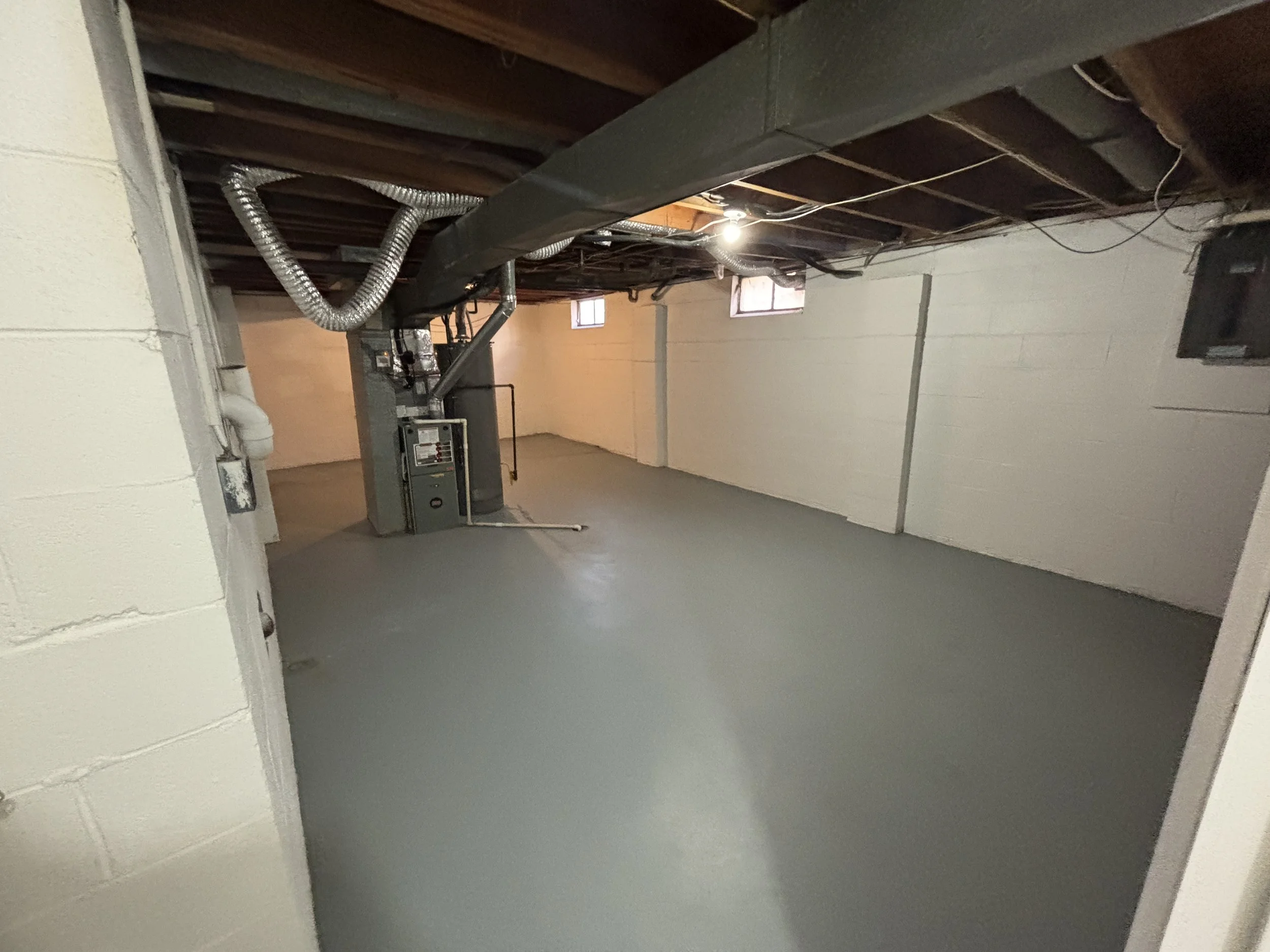 Empty basement with concrete floor, exposed wooden ceiling beams, small windows near the ceiling, and HVAC ductwork and equipment along one wall.