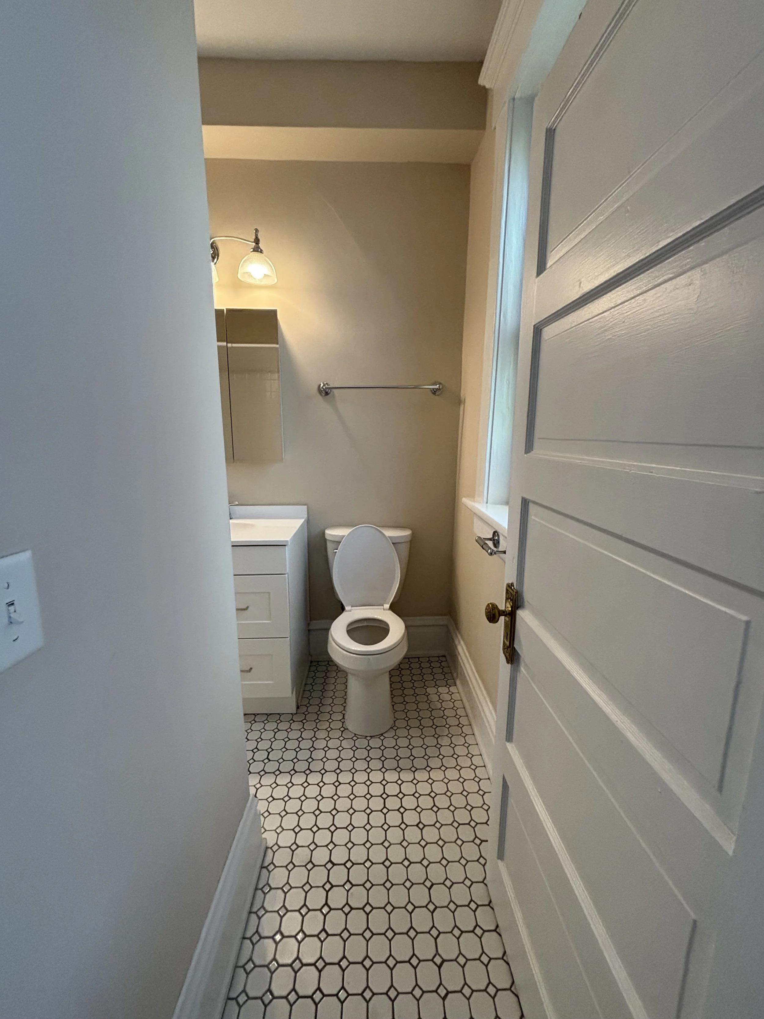 Small bathroom with a white door open, featuring a toilet, a white sink with cabinet, a small mirror, a towel bar, tile flooring, and a window on the right.