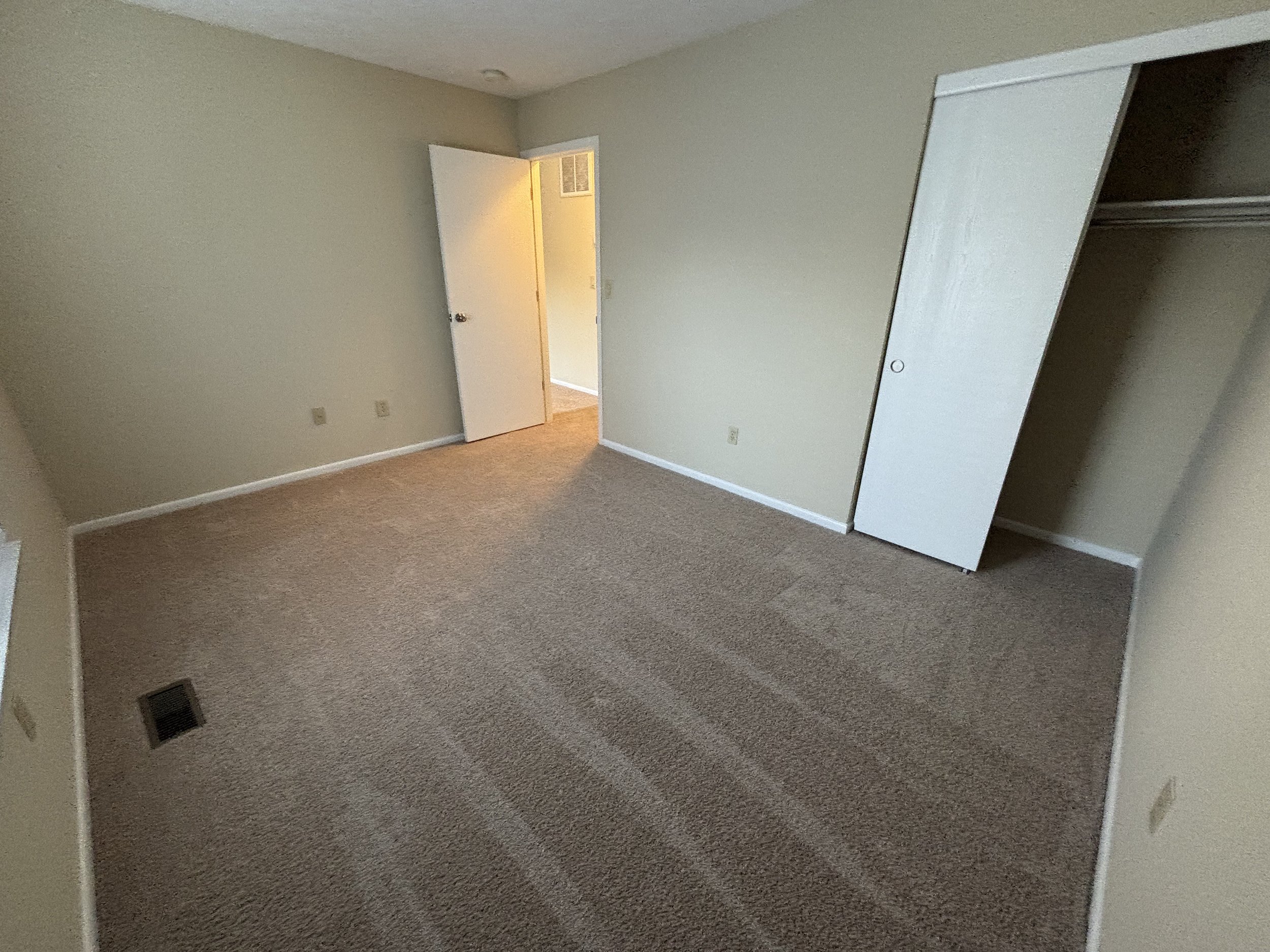 Empty bedroom with beige walls, carpeted flooring, a closet with sliding doors, and an open door leading to a hallway.