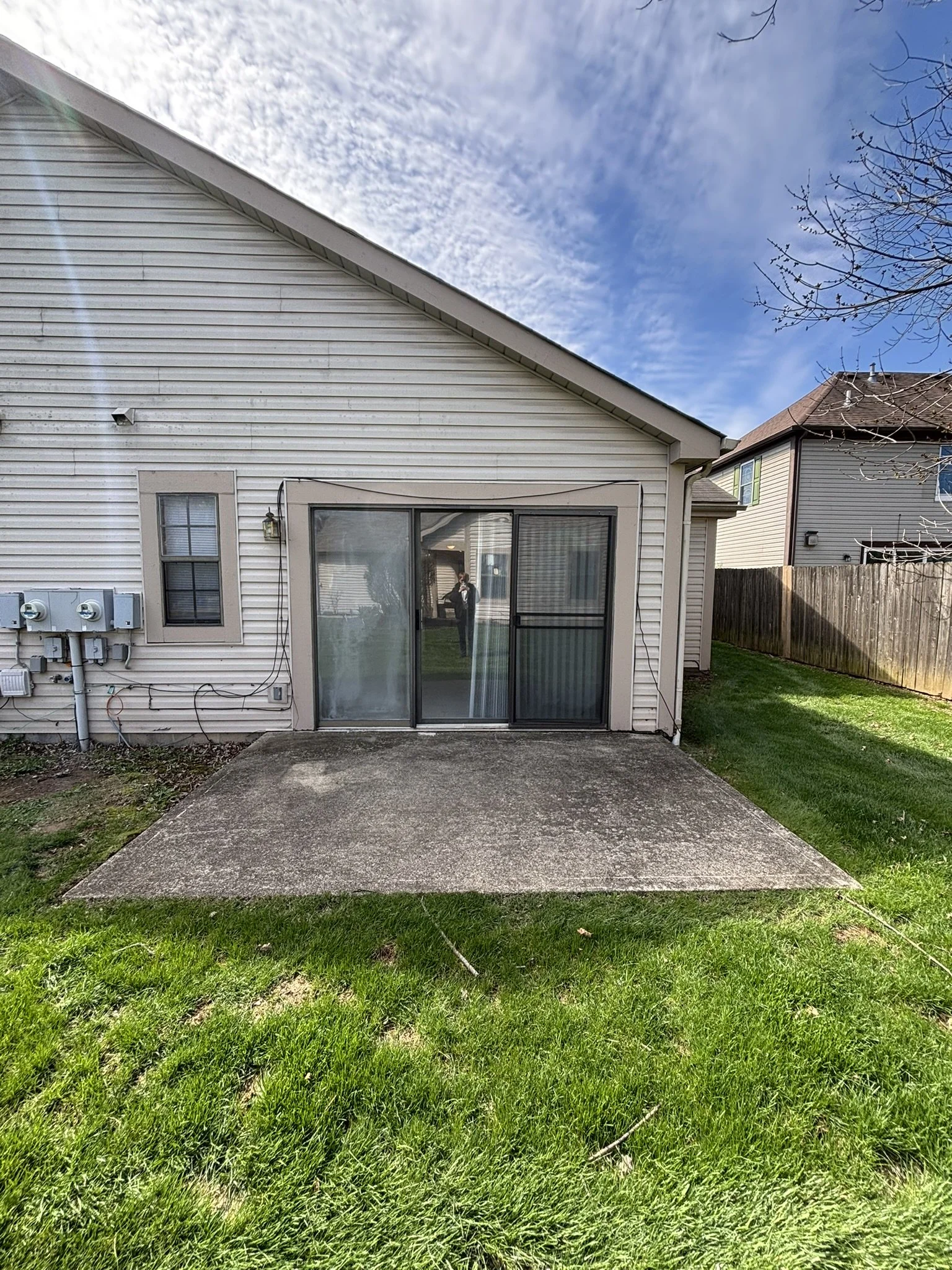 Backyard patio with concrete slab, sliding glass door, and white house exterior with two small windows and electrical meters, adjacent to a grassy lawn, with neighboring houses visible in the background and a partly cloudy sky overhead.