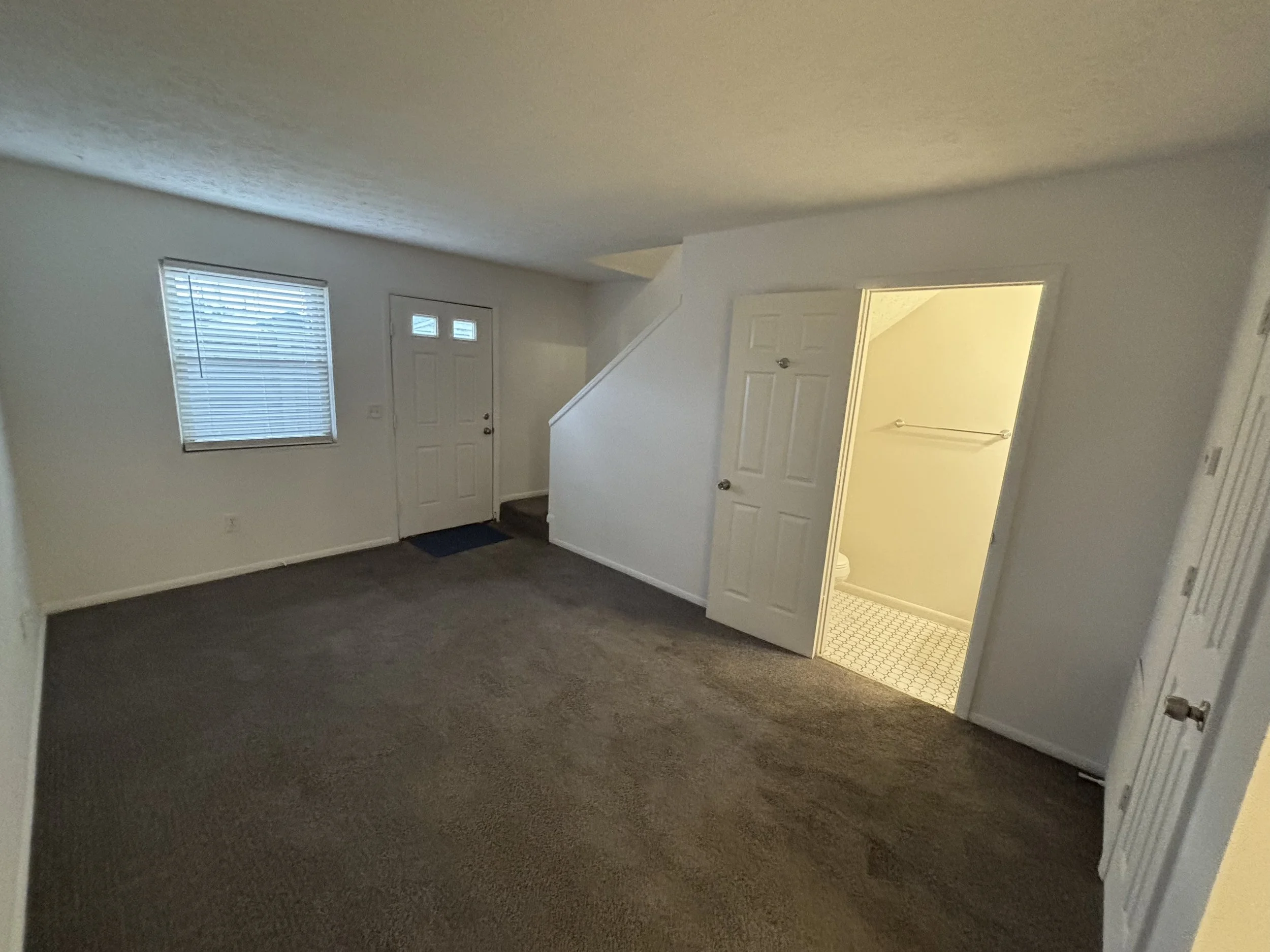 Empty living room with beige carpet, white walls, a window with blinds, a front door, and a doorway leading to a bathroom.