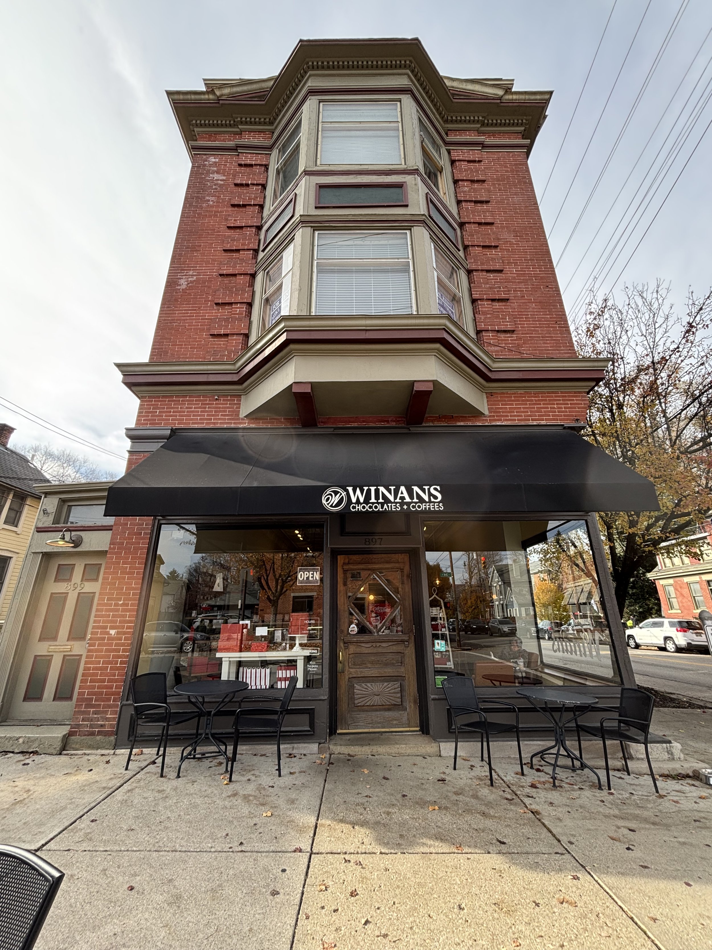 Facade of a three-story brick building with a café named 'Winans Chocolates + Coffees' on the ground floor, featuring outdoor seating with black chairs and tables, and a black awning over the entrance.