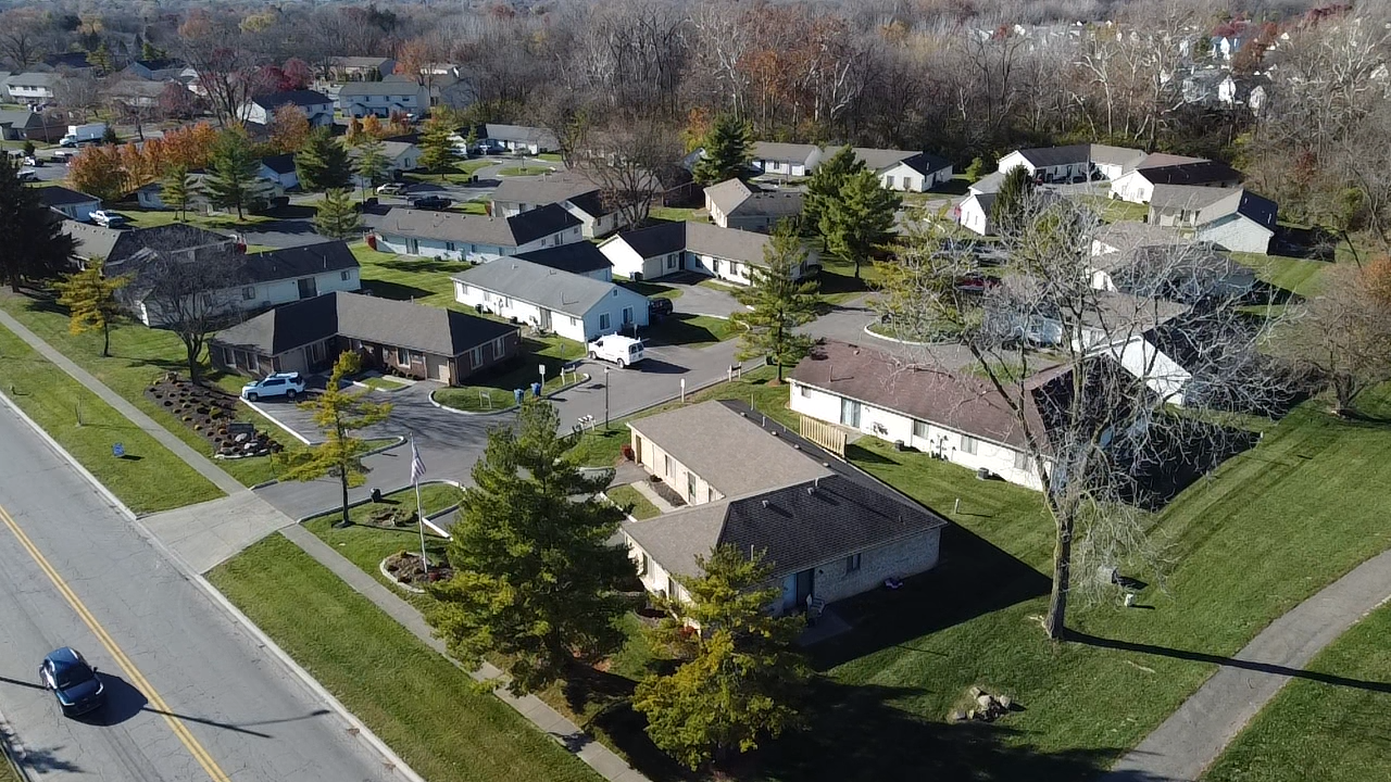 Aerial view of a small residential neighborhood with single-story houses, green lawns, trees, and a road with cars on it.