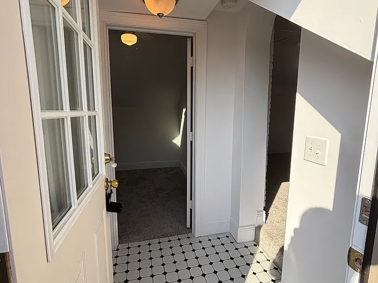 View from the front door into a small hallway with a door leading to a carpeted room, a light fixture on the ceiling, and an electrical outlet on the wall. The floor at the entrance has black and white hexagonal tiles.