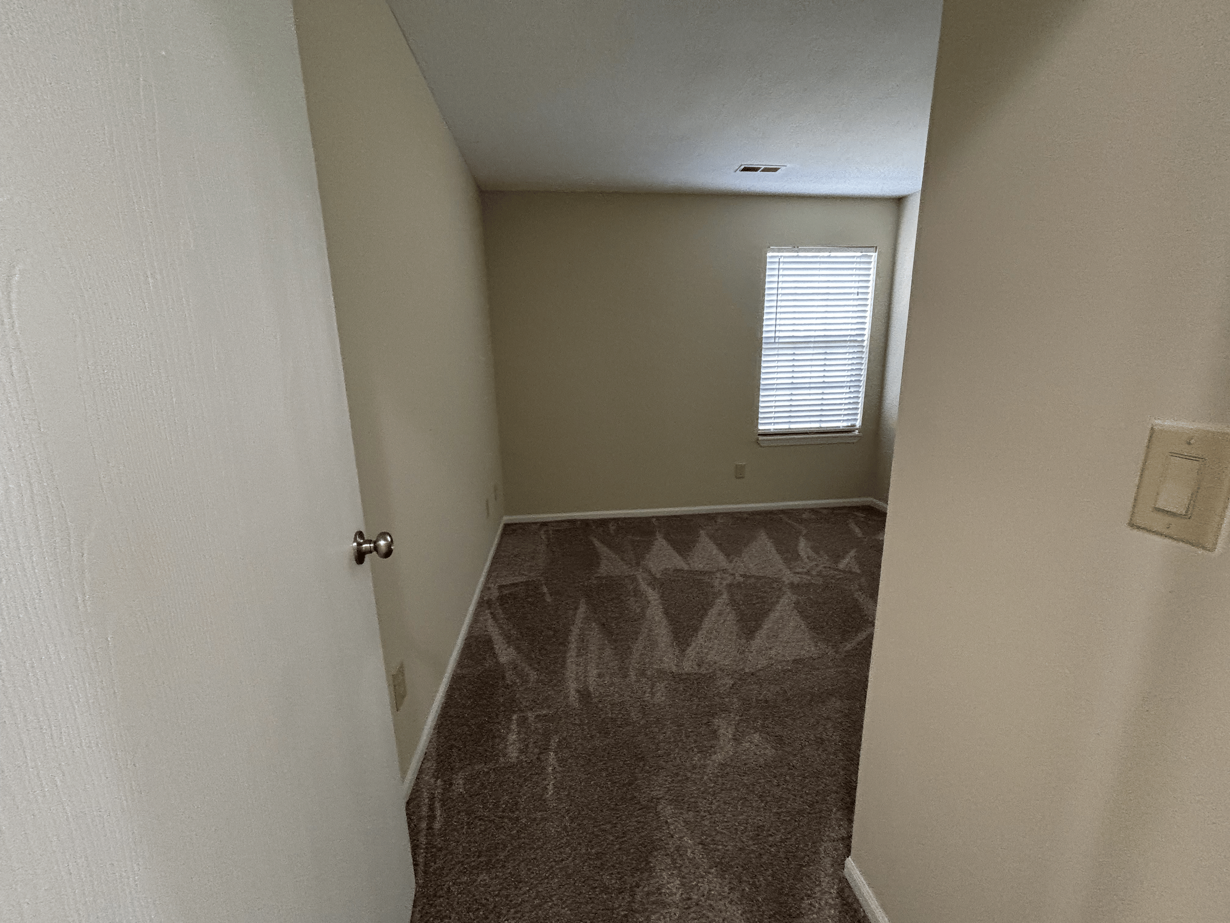 Empty room with beige walls, a window with closed blinds, and brown carpeted floor.