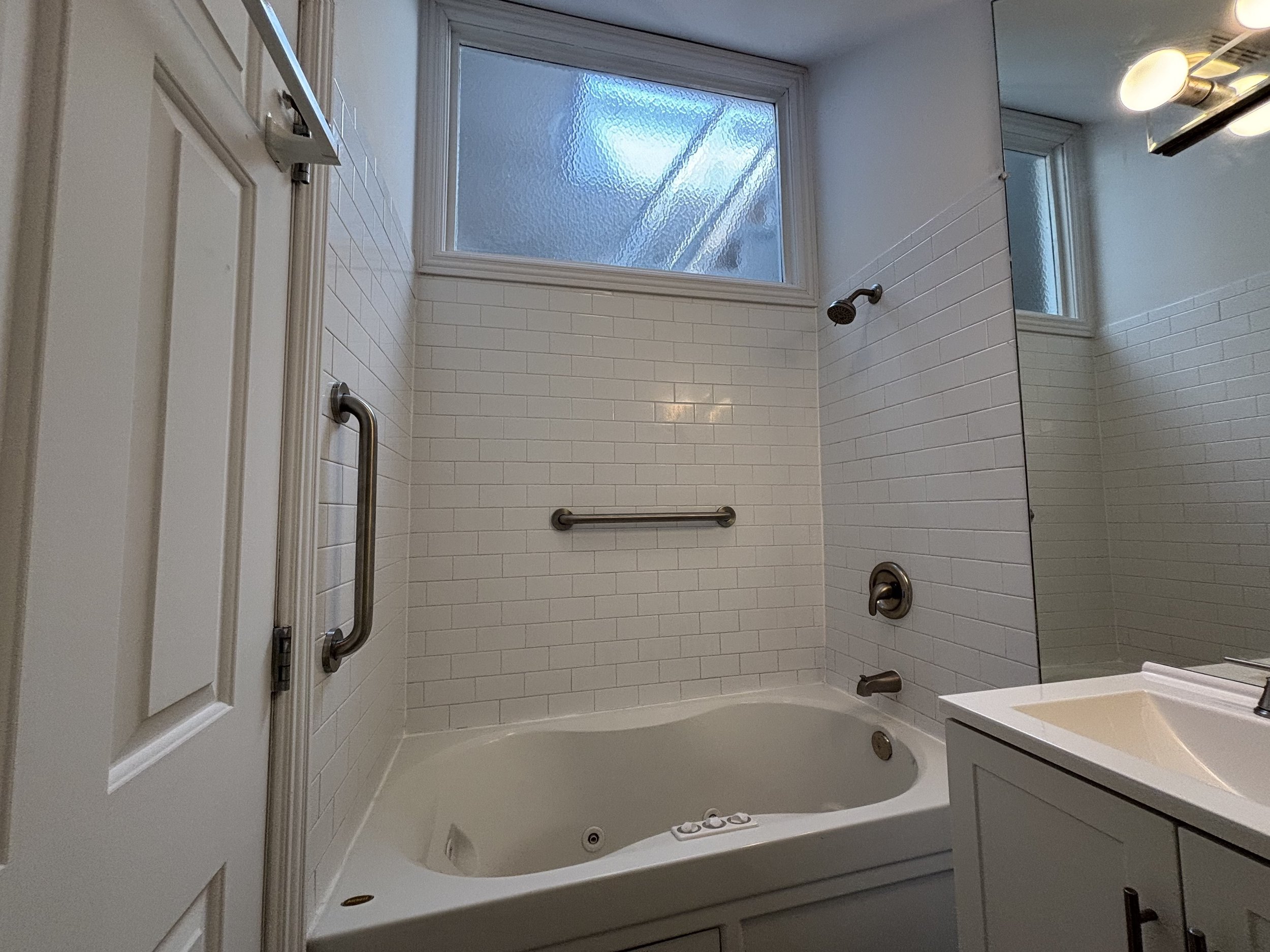 A bathroom with a white bathtub, a tiled wall, a frosted window above, a grab bar, a showerhead, a faucet, and a mirror.