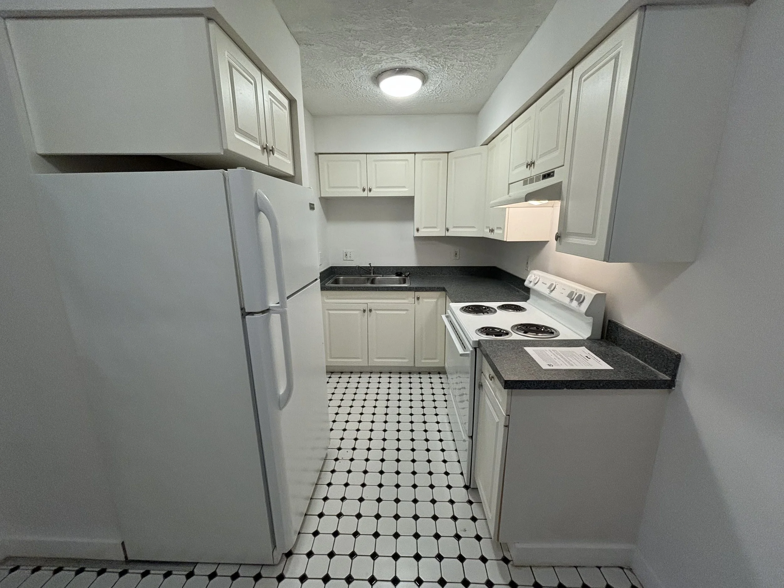 A small kitchen with white cabinets, a white refrigerator, a white stove, and a black countertop. The floor is tiled with a black and white pattern. There is a ceiling light fixture.