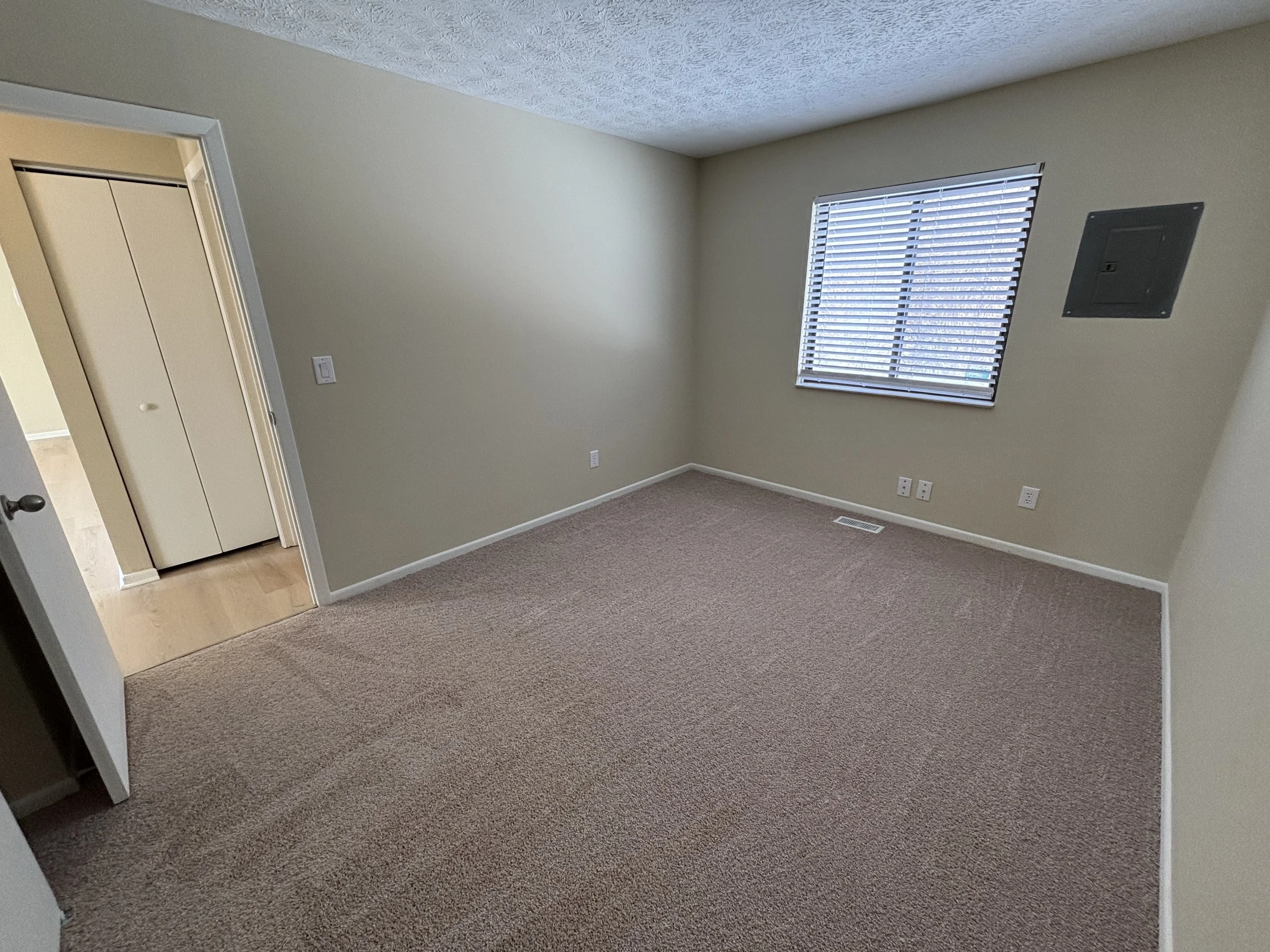 Empty beige room with window blinds, closet, and wall electrical panel, beige carpeted floor, and off-white walls.