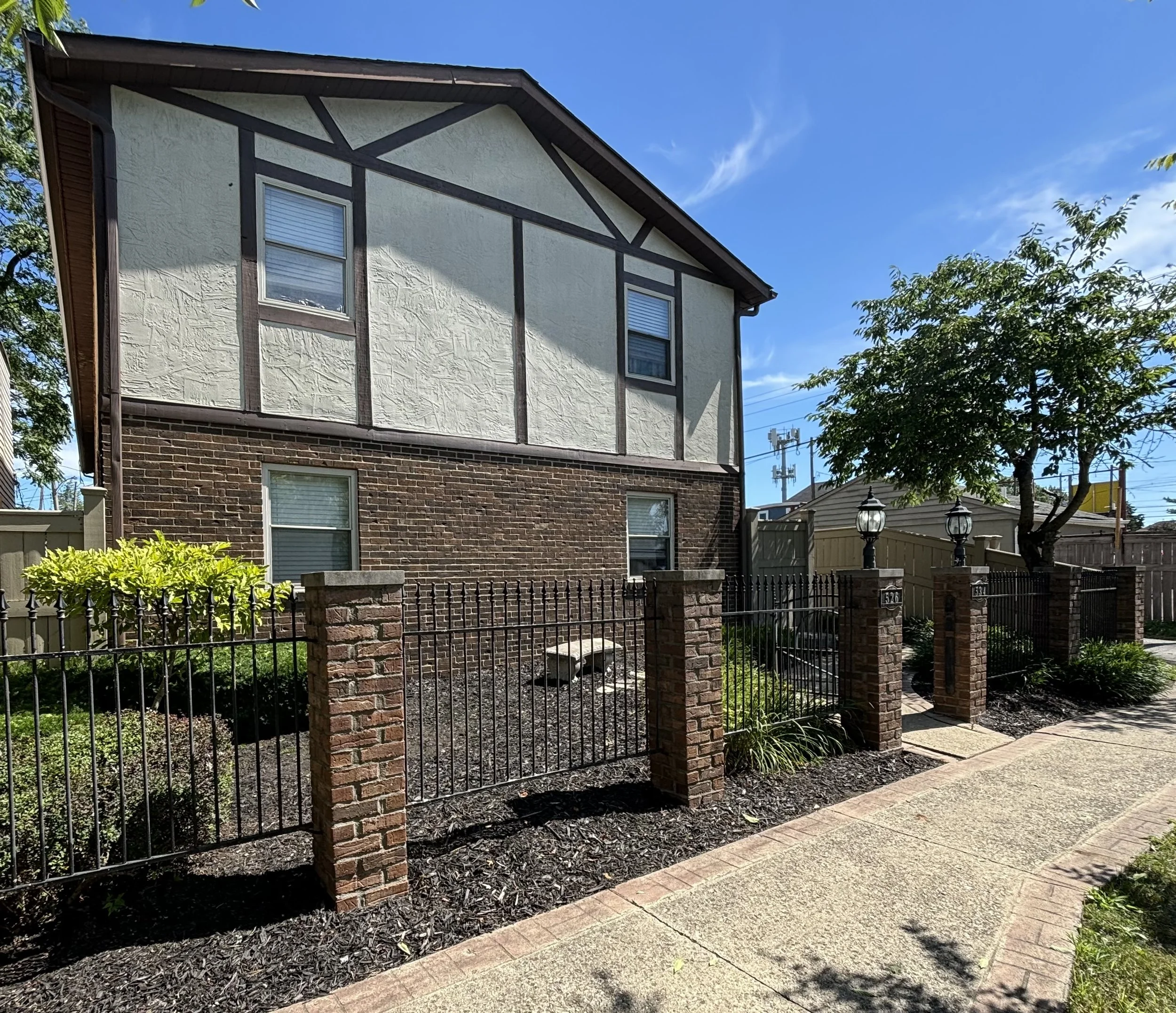 A two-story house with a brick lower level, a stucco upper level with dark wood trim, and two windows. There is a small garden with greenery and a brick and metal fence with brick columns and black lampposts along the sidewalk. A tree with green leaves is on the right side, and the sky is clear with a few clouds.