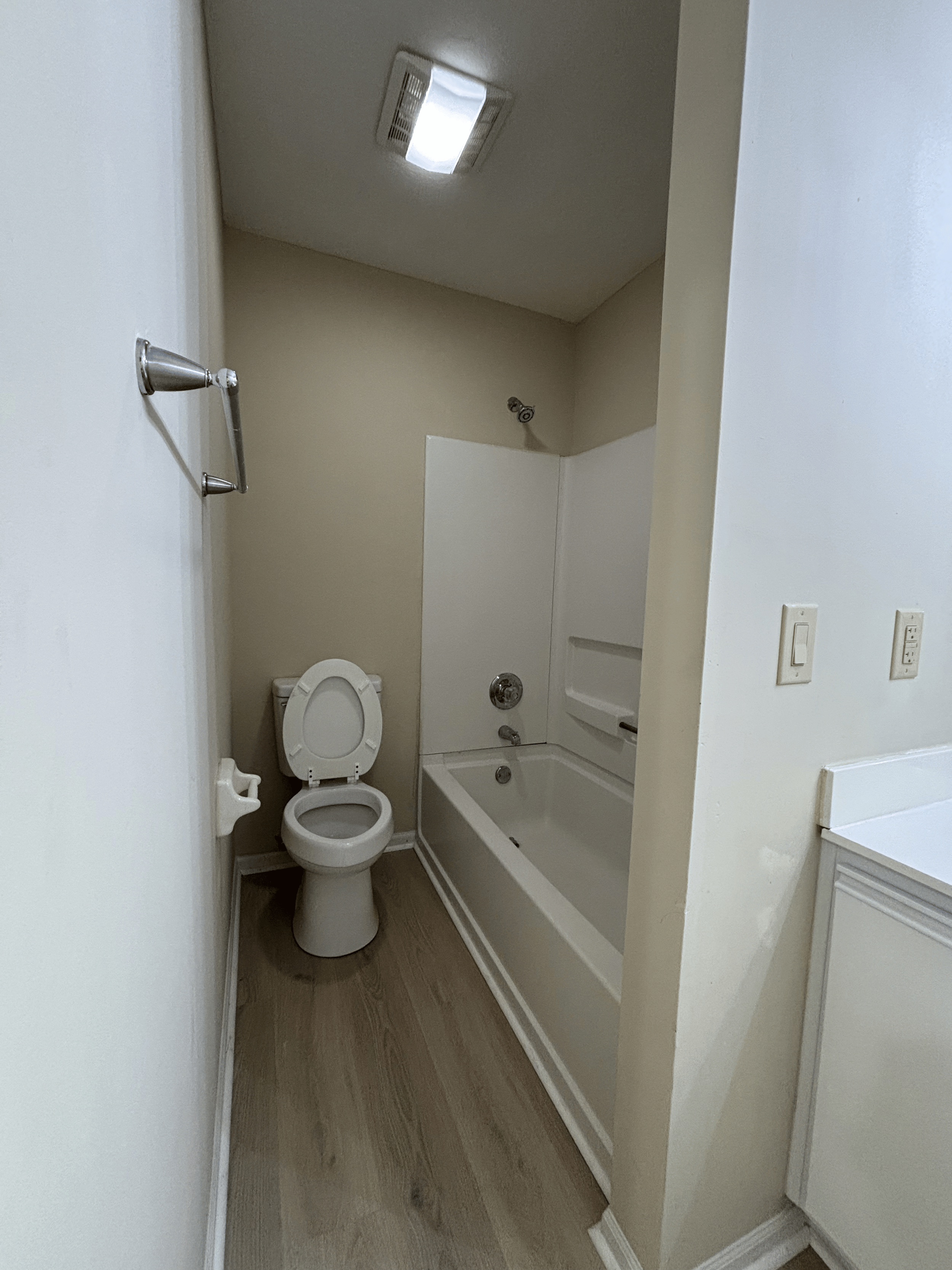 Small bathroom with a toilet and a bathtub with two shelves, beige walls, wood flooring, white trim, and a ceiling light.