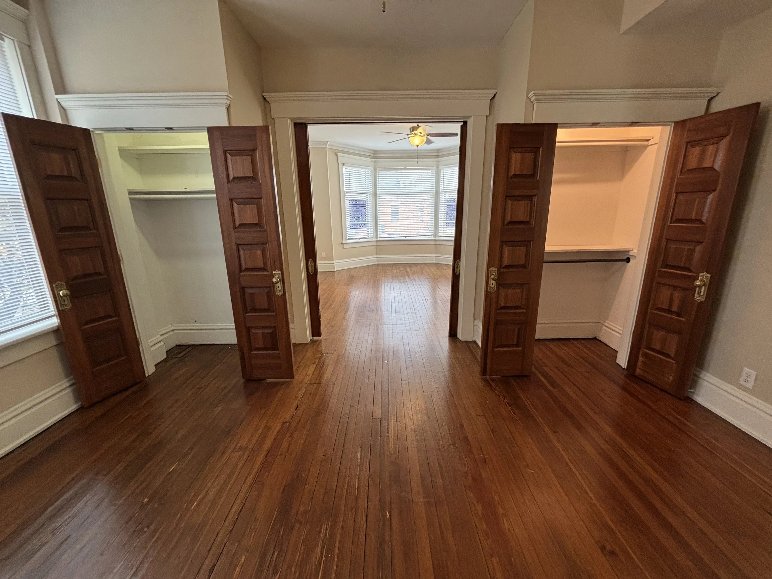 Empty bedroom with hardwood floors, two open closets with wooden doors, and a view into a bright room with large windows and a ceiling fan.