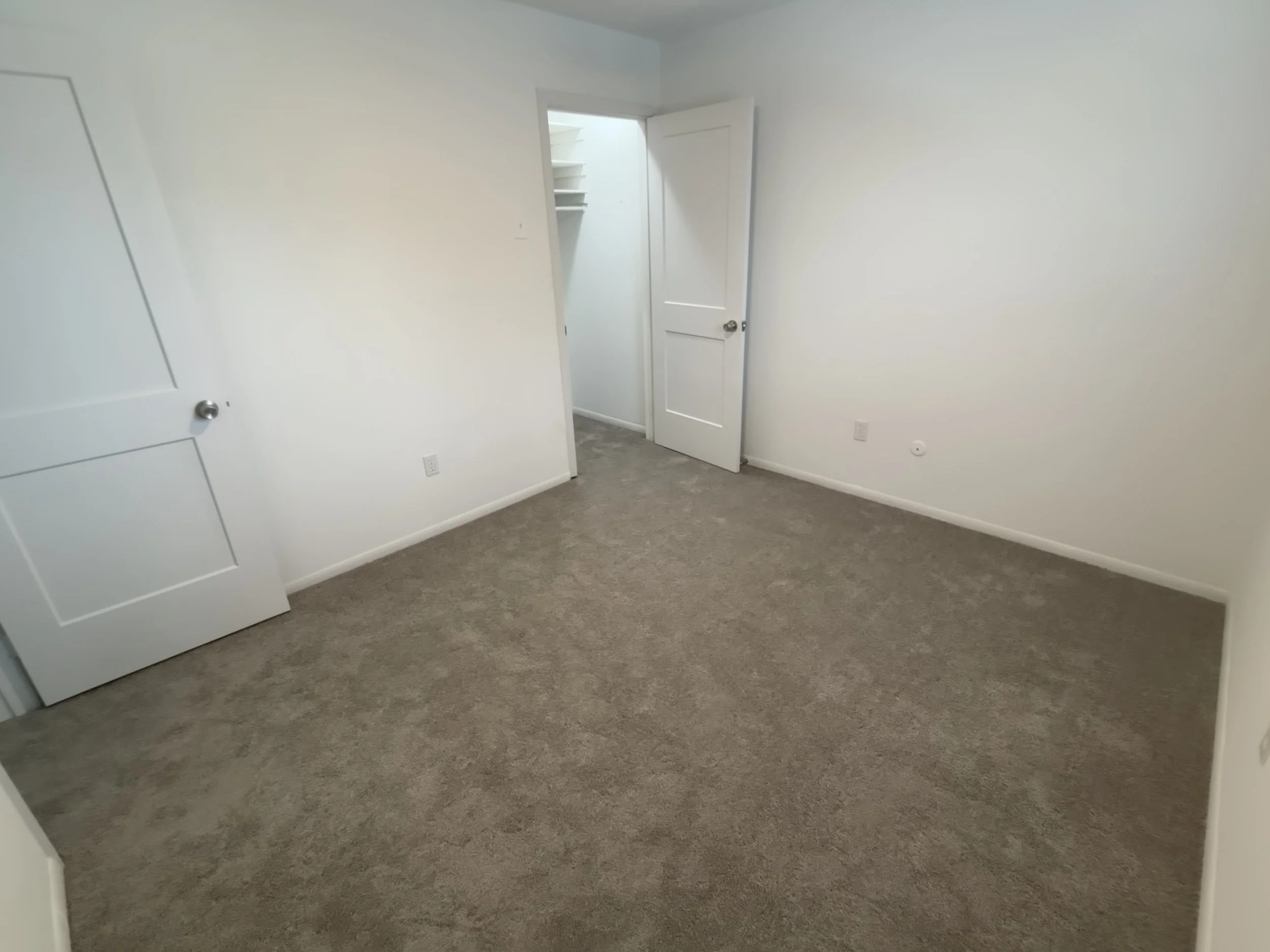 Empty room with white walls, gray carpet, open door, and closet with shelves.