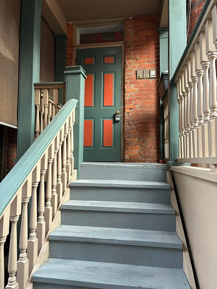 A front porch with gray wooden stairs leading up to a green door with orange panels, flanked by wooden railings painted in light beige and green. To the right of the door is a red brick wall with a small window above and a mailbox.