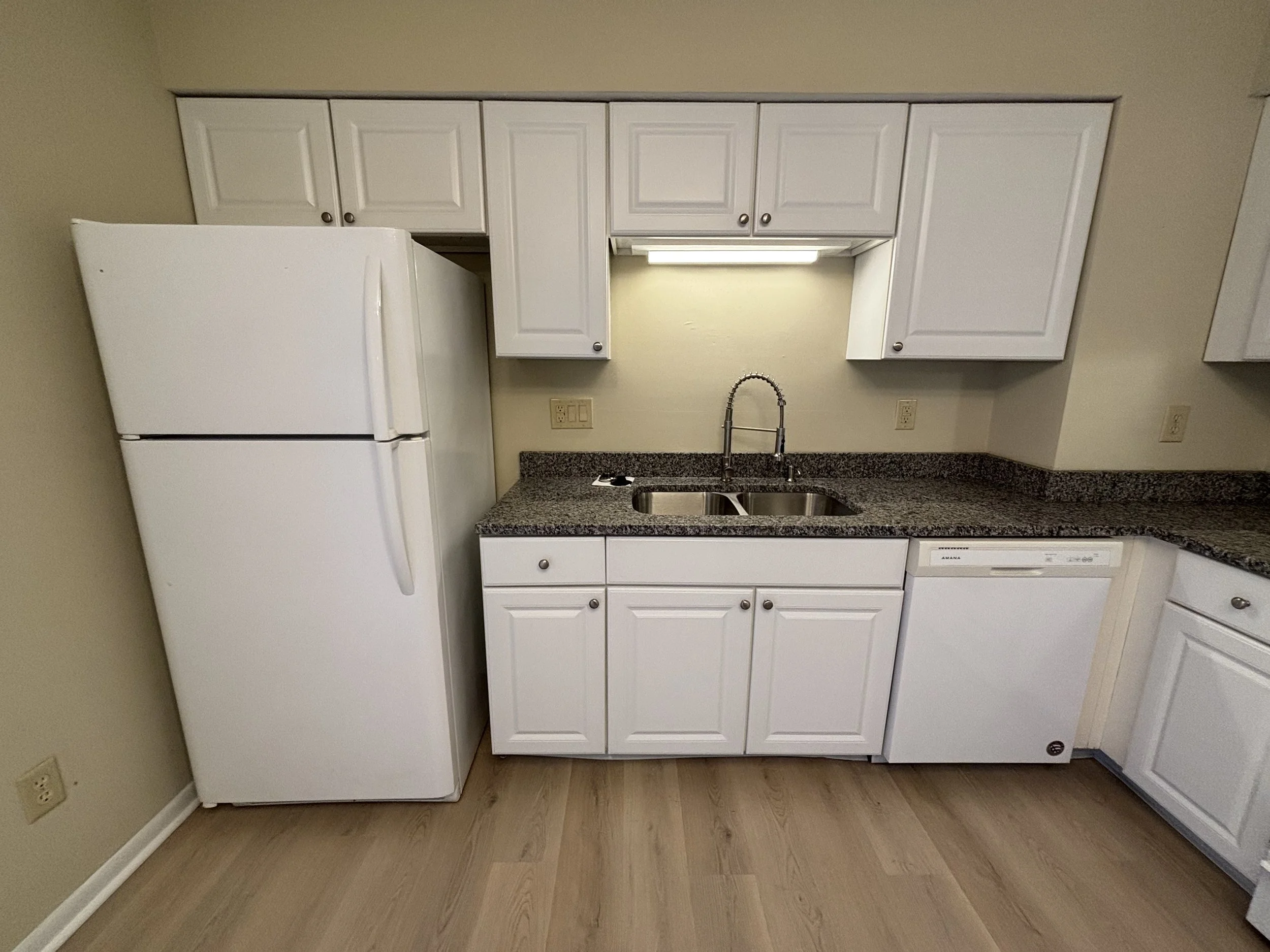 A kitchen with white cabinets, a granite countertop, a double sink with a high-arc faucet, a white refrigerator, and a dishwasher under the counter. The floor is wood, and walls are beige.