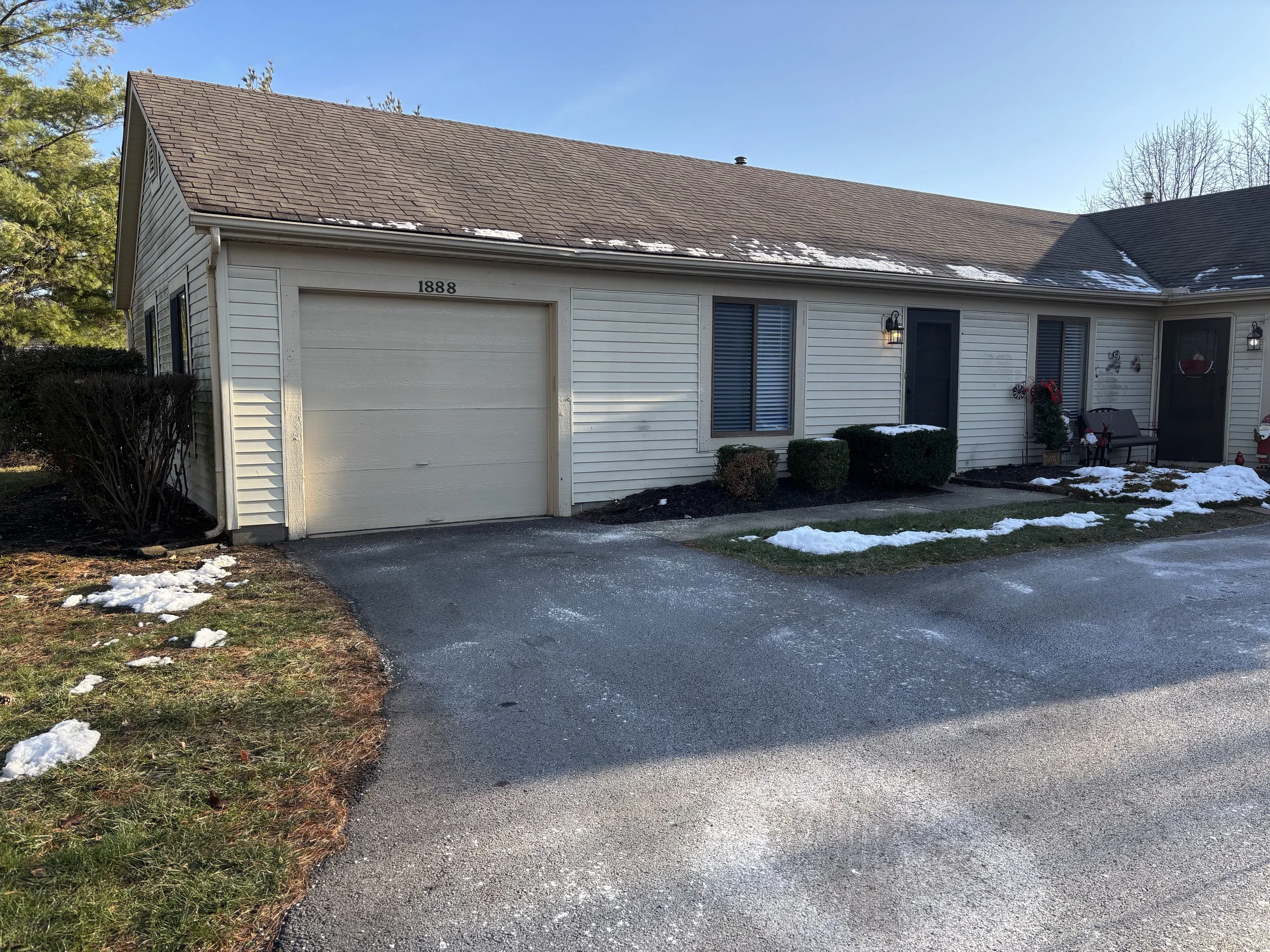 Front view of a white house with a one-car garage, snow patches on the roof and small patches on the yard, and holiday decorations outside.