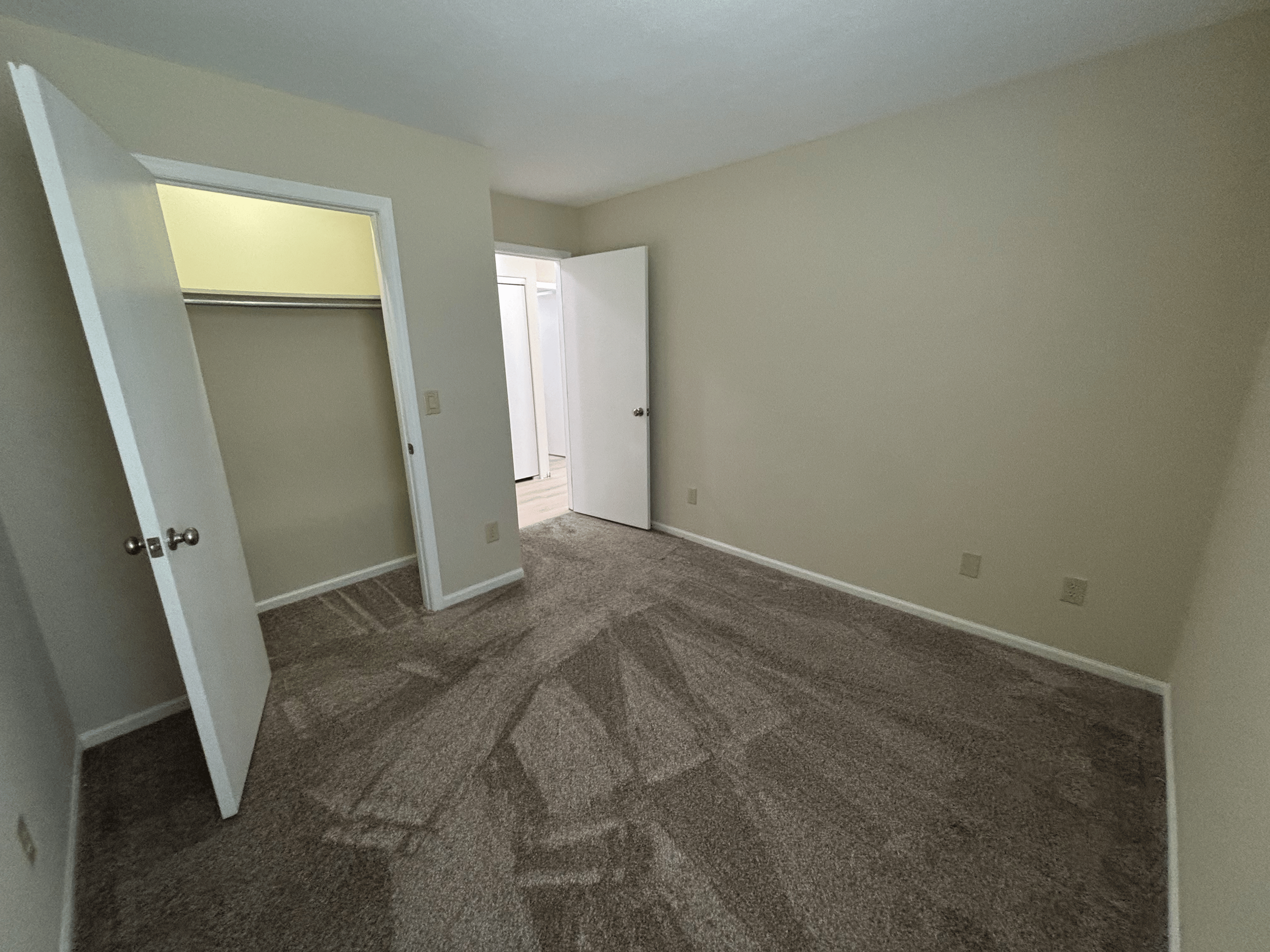 Empty beige bedroom with open closet door revealing an empty closet and a second open door leading to a hallway or bathroom.