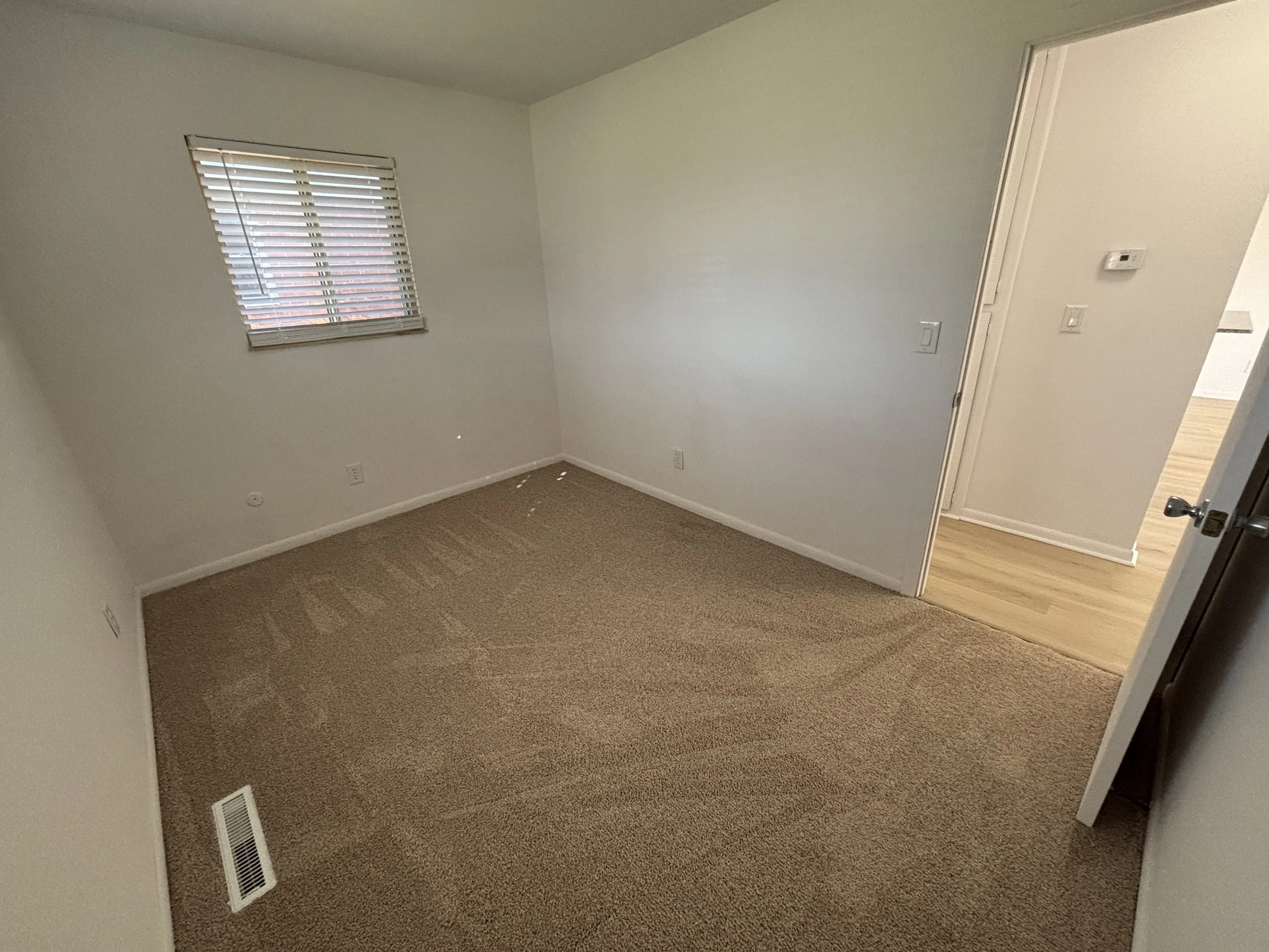 Empty room with beige carpet, white walls, a window with blinds, and a doorway leading to another room with light wood flooring.