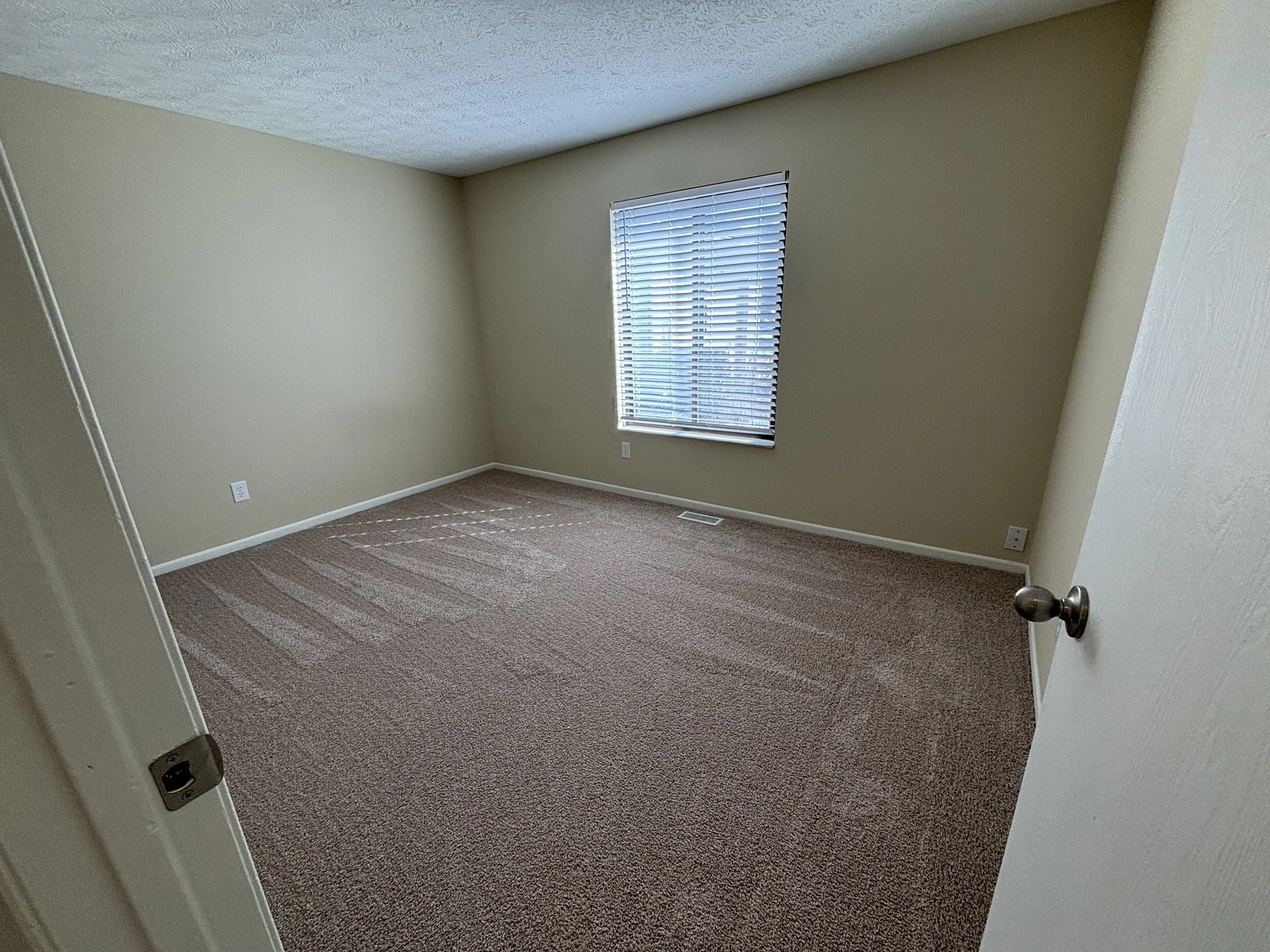 Empty room with beige walls, tan carpet, a window with blinds, and a ceiling vent.