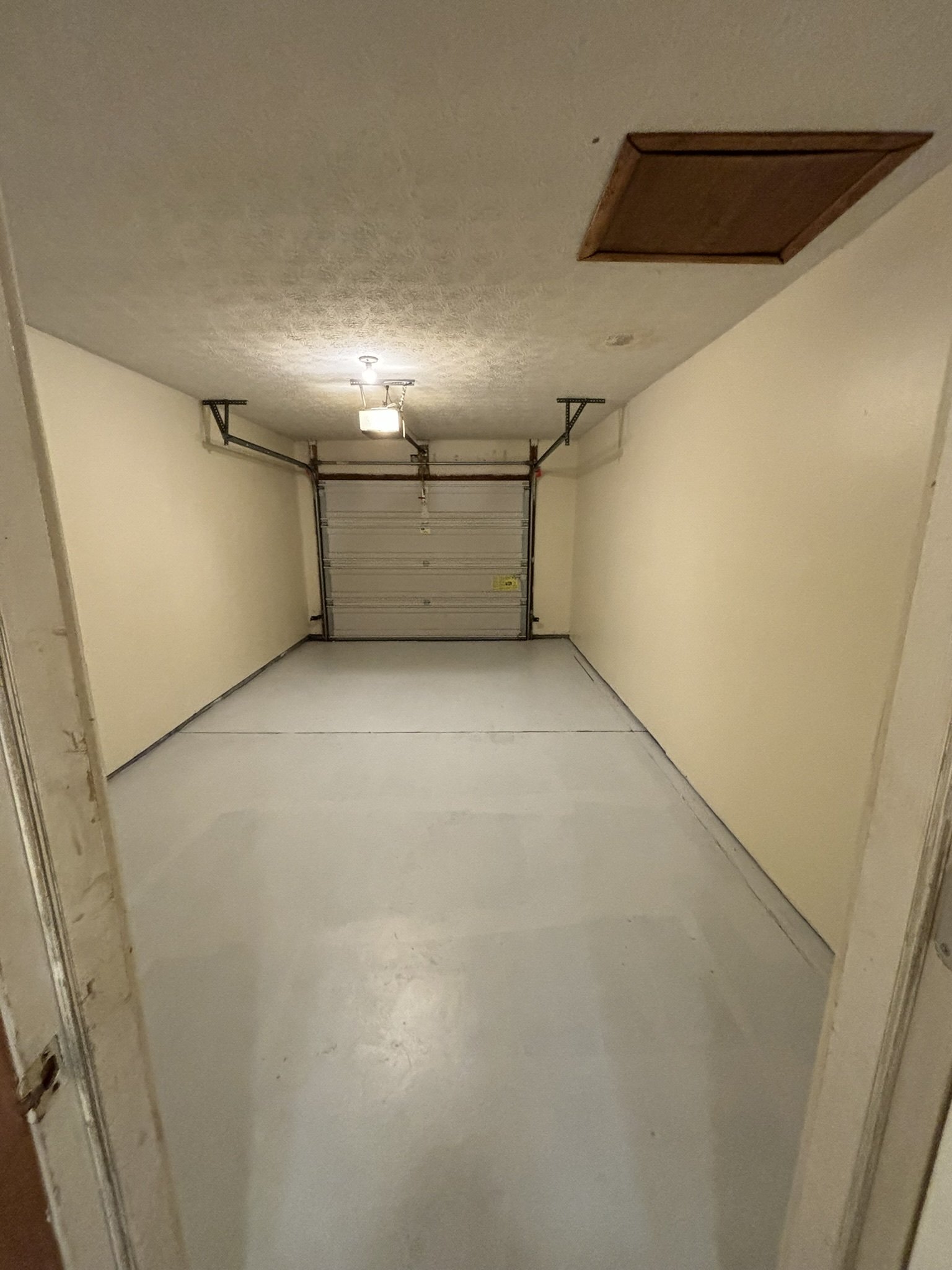 Empty garage with white floor, beige walls, and a closed garage door, interior with ceiling light, access panel on ceiling