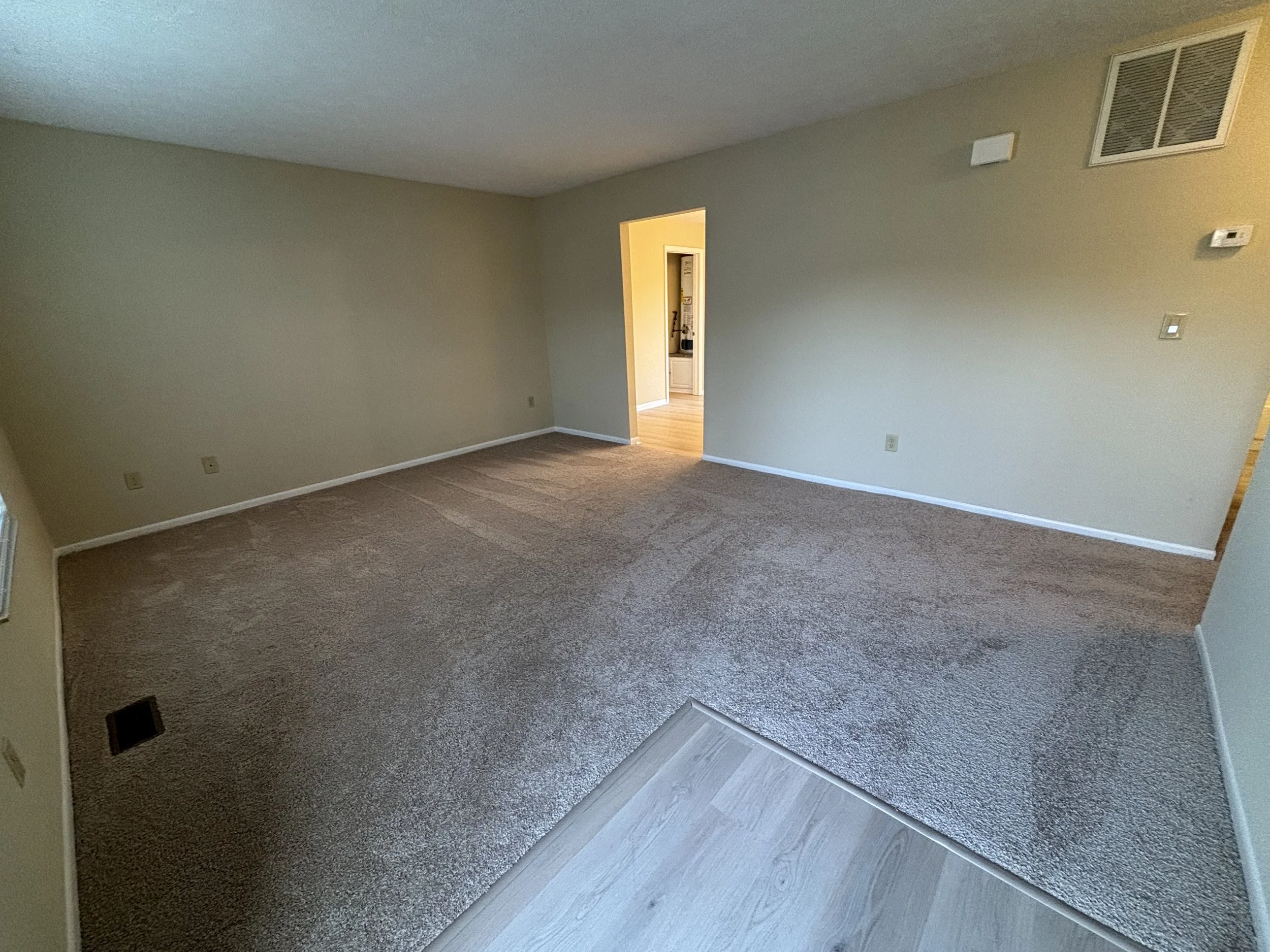 Empty living room with beige carpet, light yellow walls, a doorway leading to another room, and a vent on the ceiling.