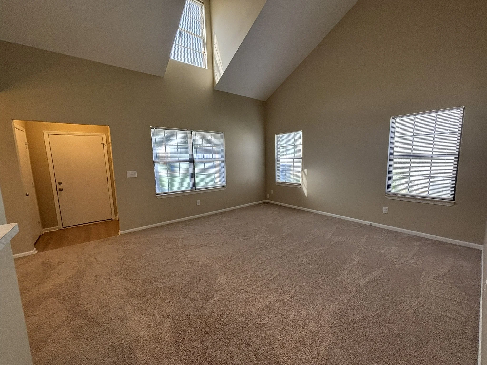 Empty living room with beige walls, large windows with blinds, carpeted floor, vaulted ceiling with skylight, and an open doorway leading to another room.