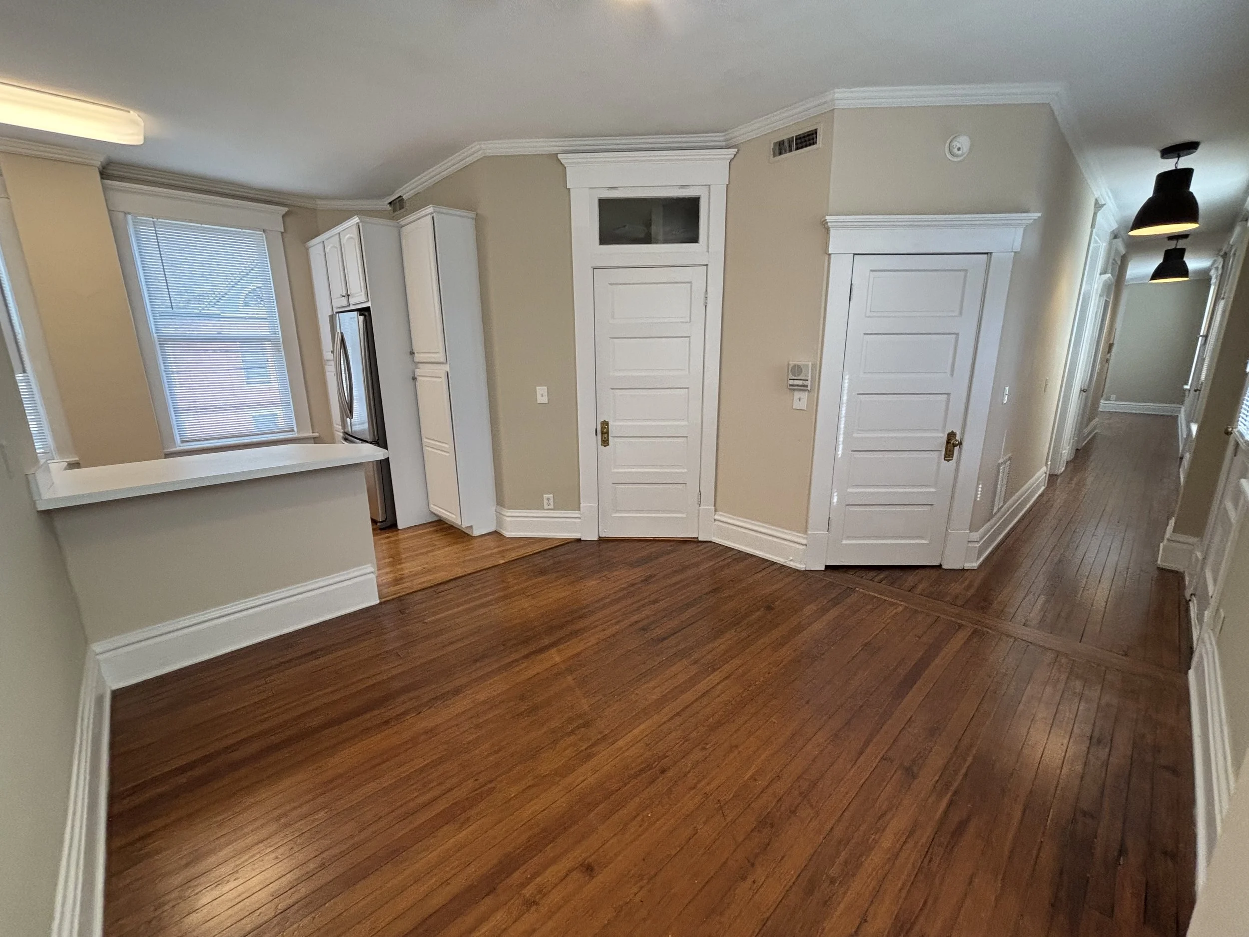 Empty room with hardwood floors and white trim, two windows with blinds, kitchen with stainless steel appliances, and hallway with black pendant lights.