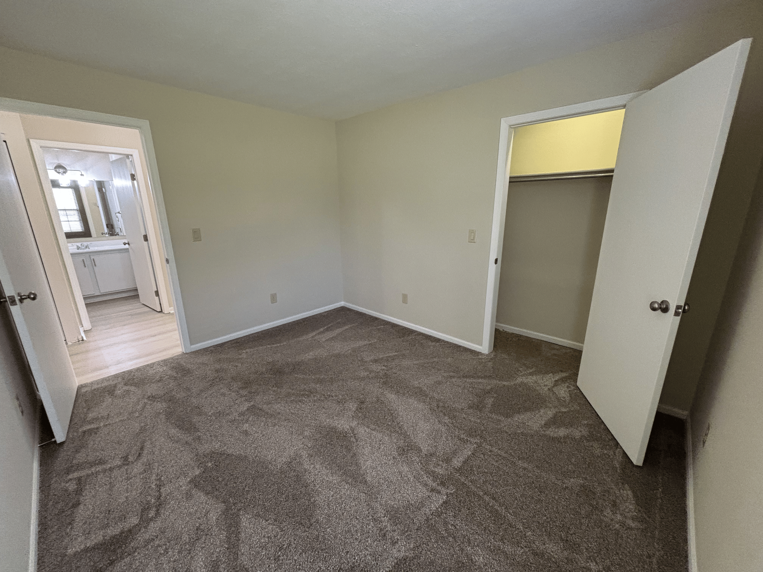 Empty bedroom with beige walls, brown carpet, open closet with a single metal rod, and doorway leading to bathroom with vanity and mirror.