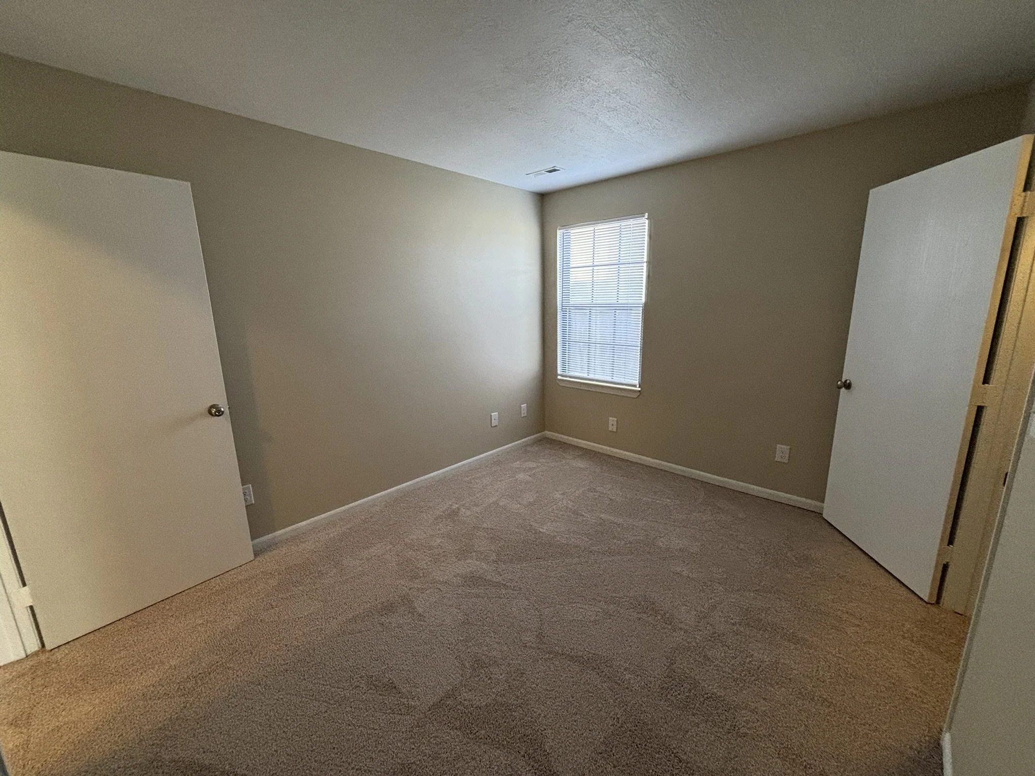 Empty bedroom with beige walls, a window with blinds, carpeted floor, and two closed doors.