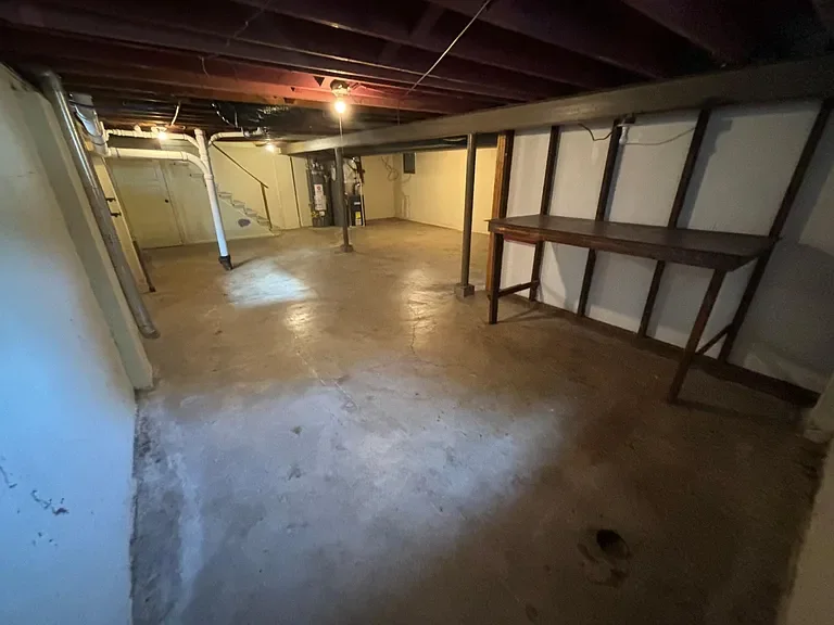 Empty basement with concrete floor, exposed ceiling beams, a wooden workbench along the wall, and pipes running along the ceiling.
