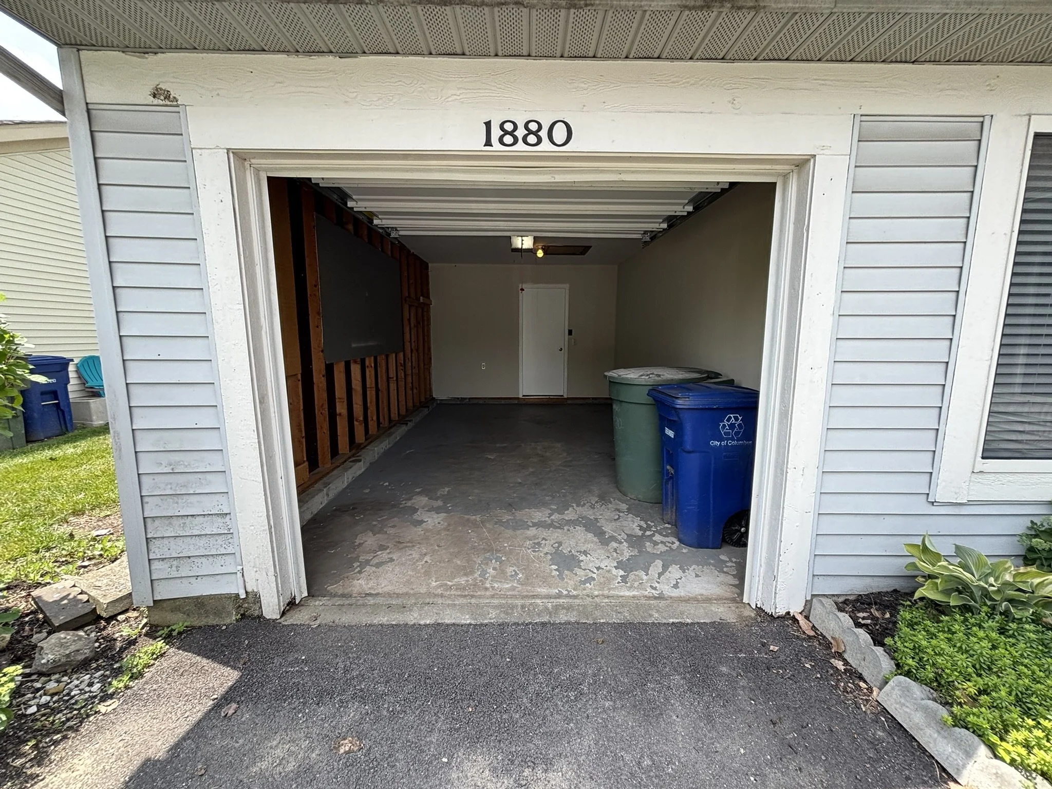 Empty garage with two trash cans, one blue and one green, and a wooden wall inside the garage. The house number 1880 is above the garage entry.