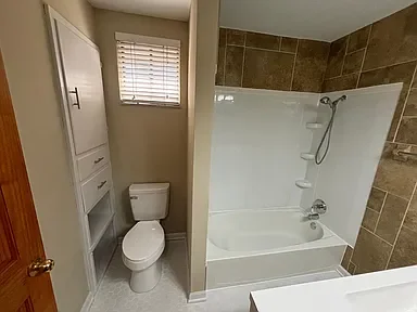 Small bathroom with a toilet, a white storage cabinet, a window with blinds, and a shower with a white surround and brown tiled wall.