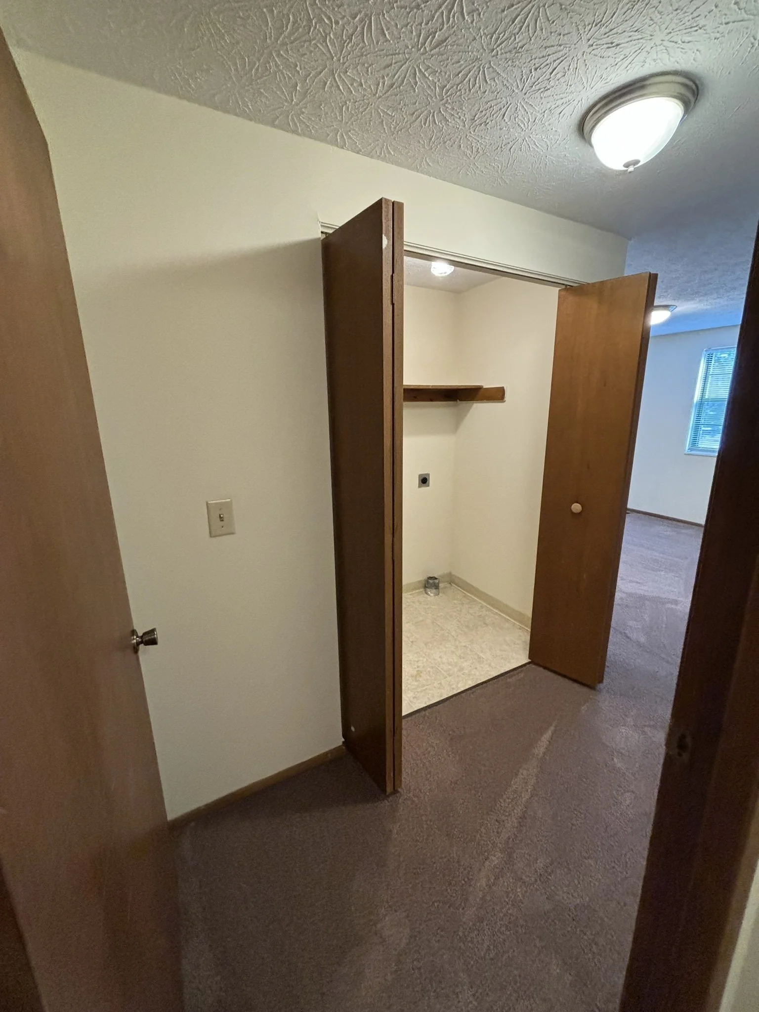 Interior view of a small closet with open wooden bi-fold doors, a wooden shelf inside, and a water supply connection on the wall, located in a room with beige carpeted flooring and textured ceiling.