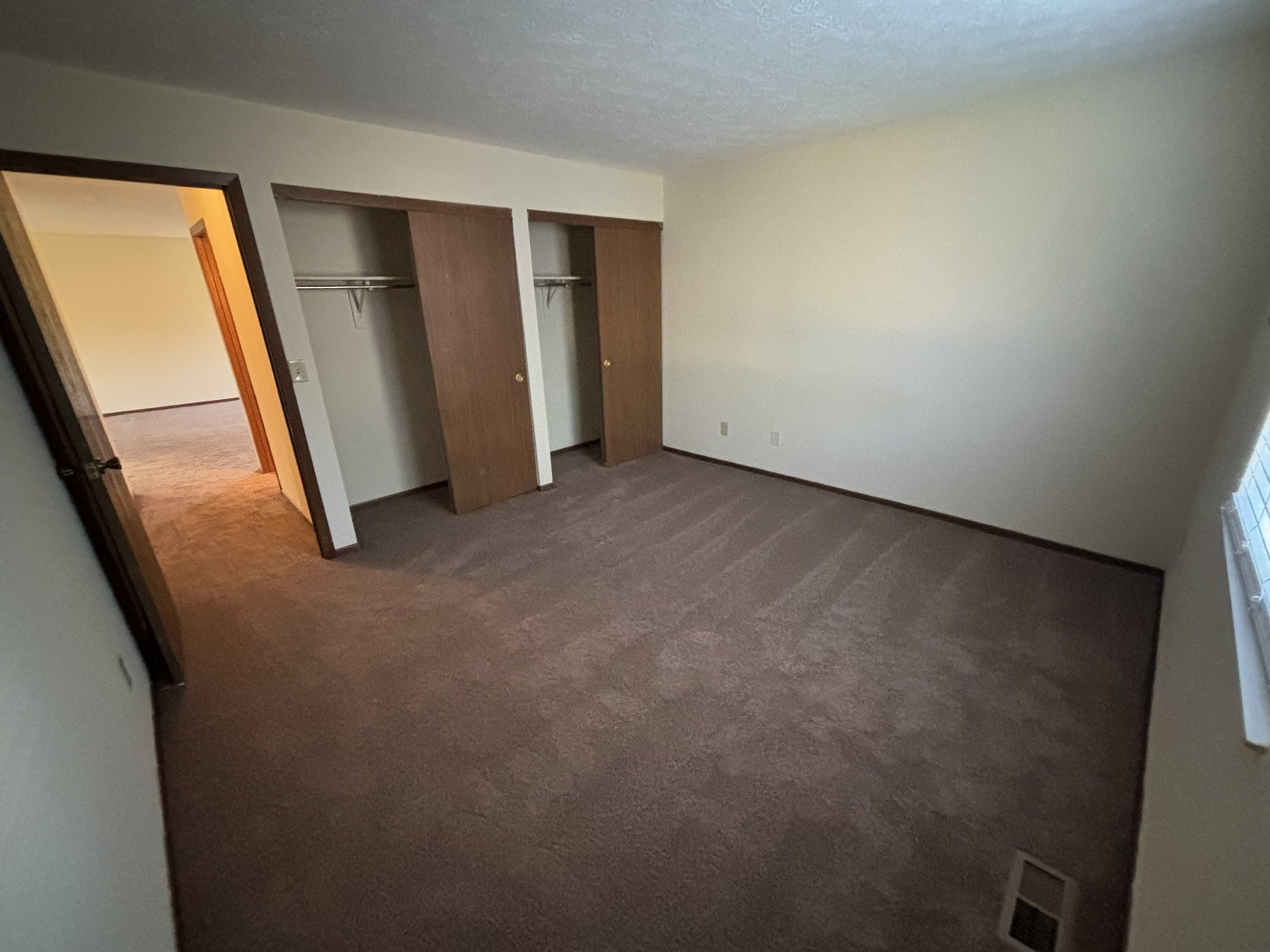 Empty bedroom with brown carpet, a window on the right, and open closet doors revealing a closet with a shelf and hanging rod.