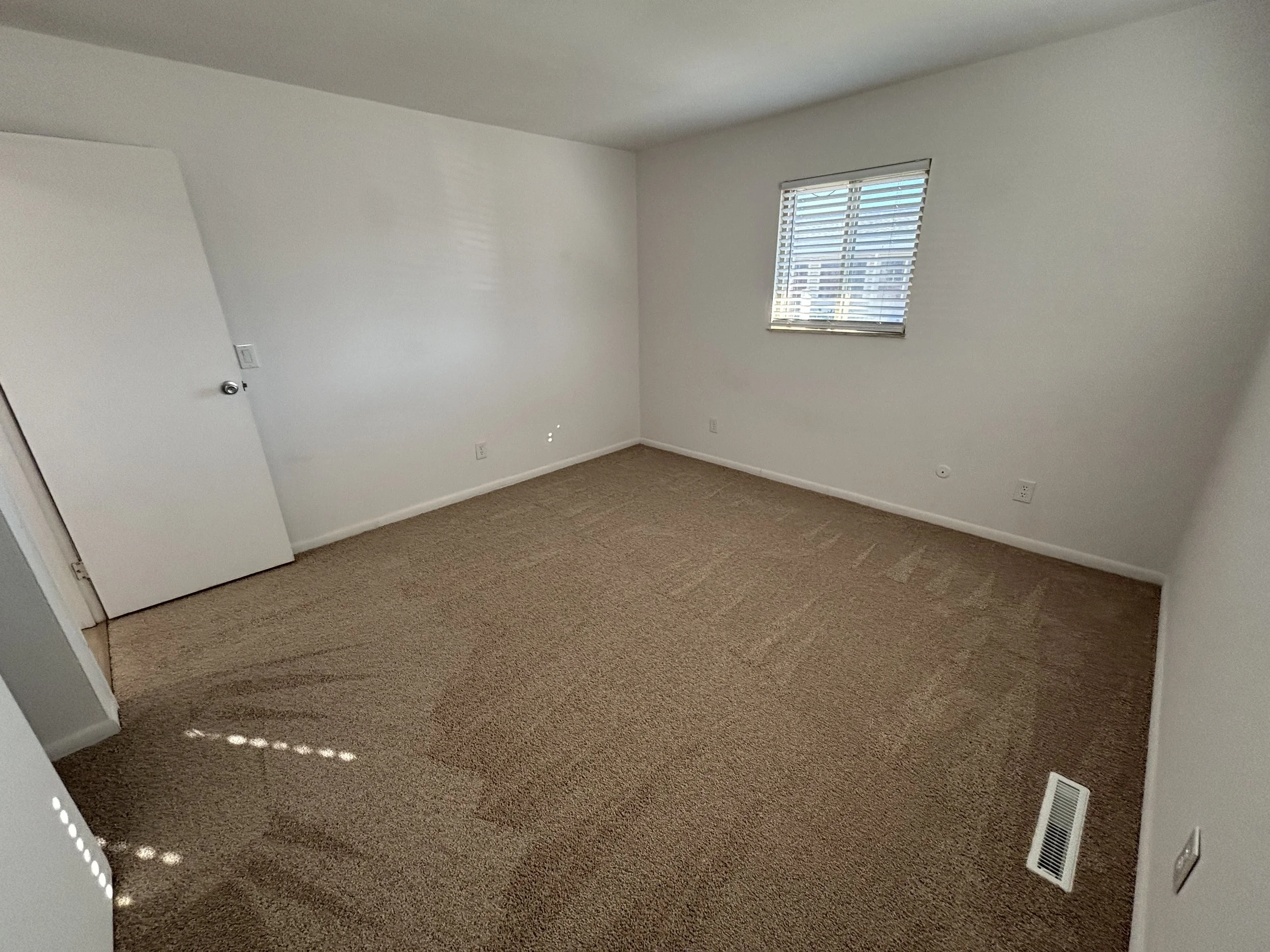 Empty room with beige carpet, white walls, a window with blinds, and a closed door.
