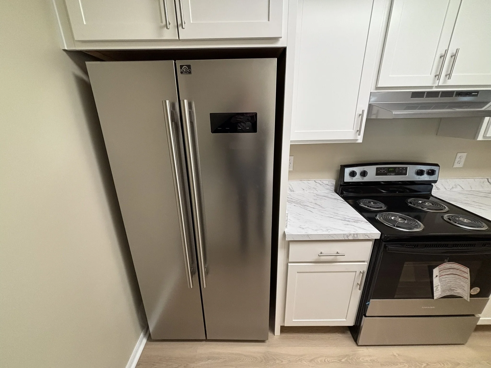 Stainless steel refrigerator next to white cabinets and a black electric stove with four burners in a kitchen with a light wood floor.