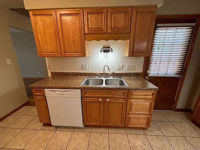 Kitchen with wooden cabinets, granite countertop, double sink, dishwasher, tiled floor, and a door with blinds.