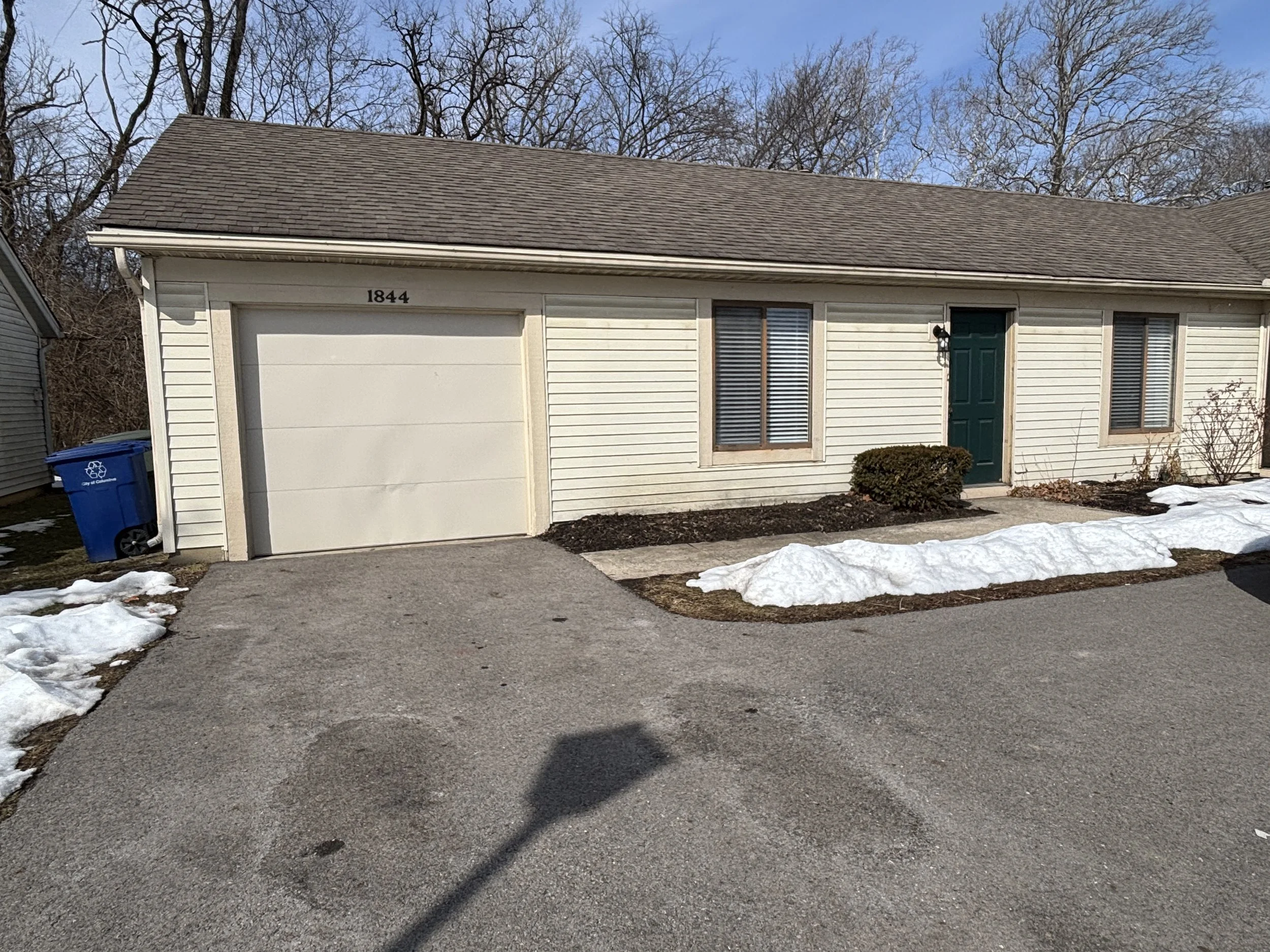 A single-story house with cream siding and a brown shingled roof, featuring a garage with a white door, a green front door, window shutters, a small bush, patches of snow on the ground, and leafless trees in the background.