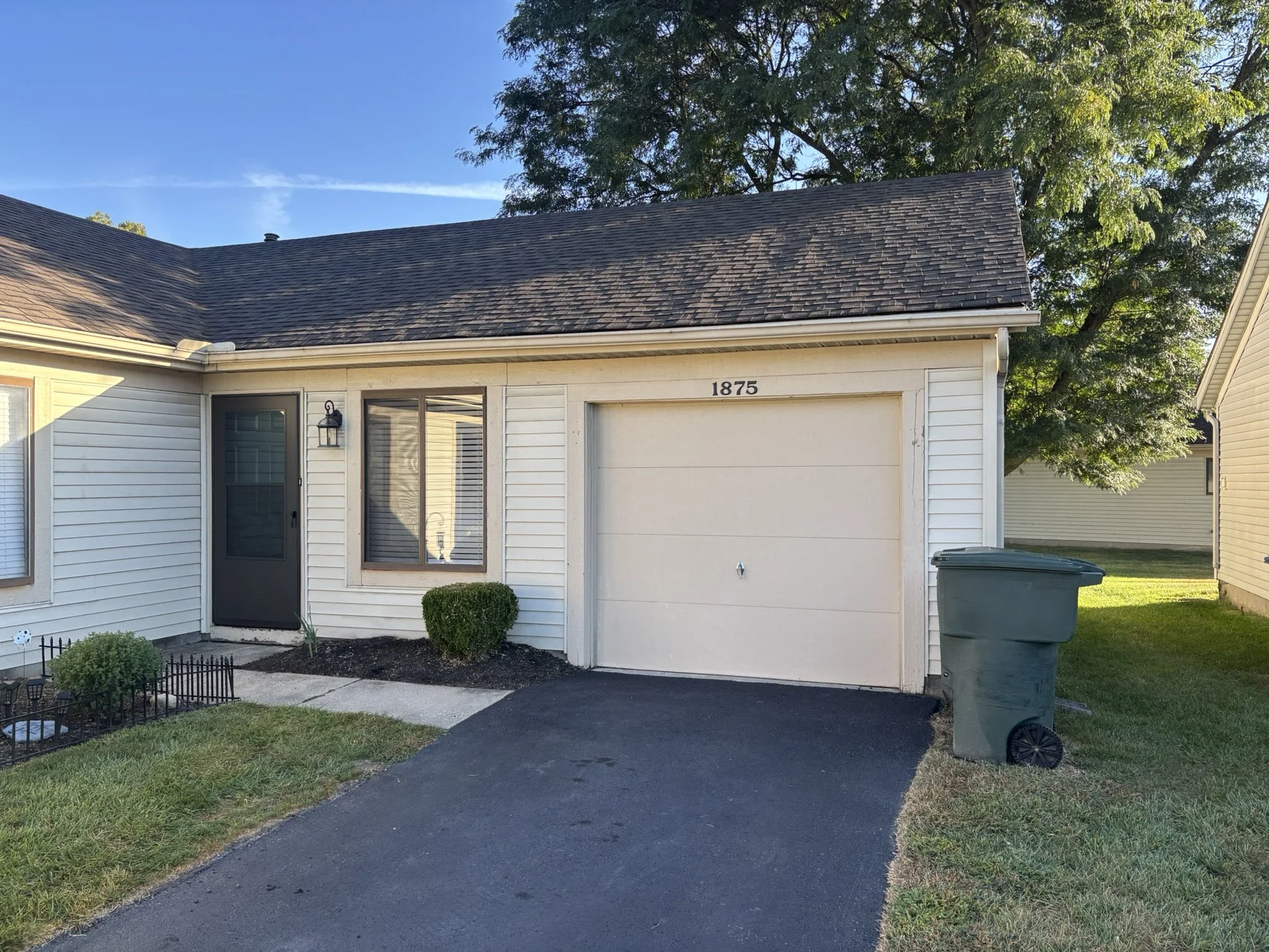 Front view of a single-story house with white siding, a black asphalt driveway, a green trash bin next to the garage, and a small landscaped area with bushes and a sidewalk. House number 1875 visible above the garage door, and a tree with green leave