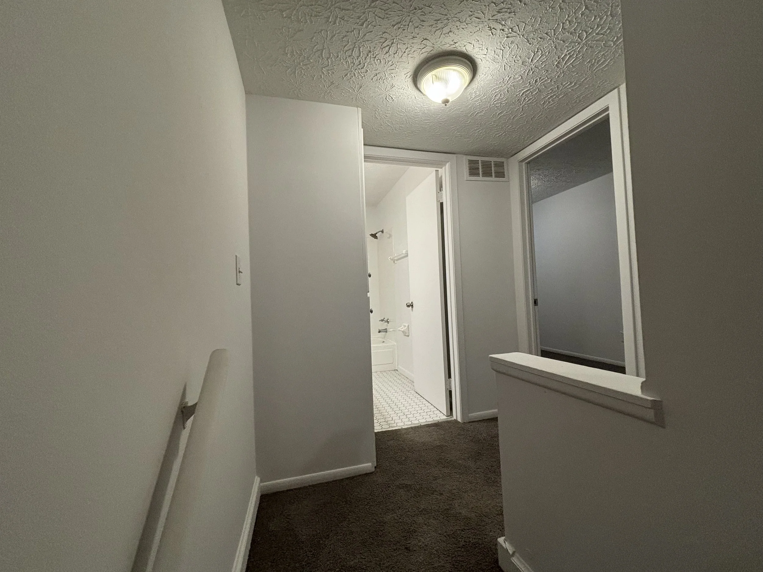 An interior view of a hallway leading to a bathroom, with a white door slightly open revealing a bathtub, seen from a slightly tilted angle with brown carpeted flooring and white walls.