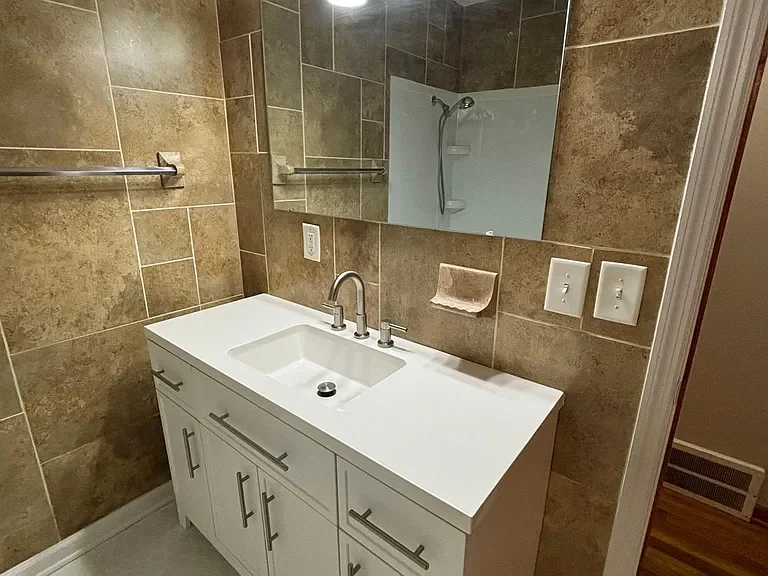 Bathroom sink with a large mirror, beige tile walls, a towel rack, and a shower reflected in the mirror.