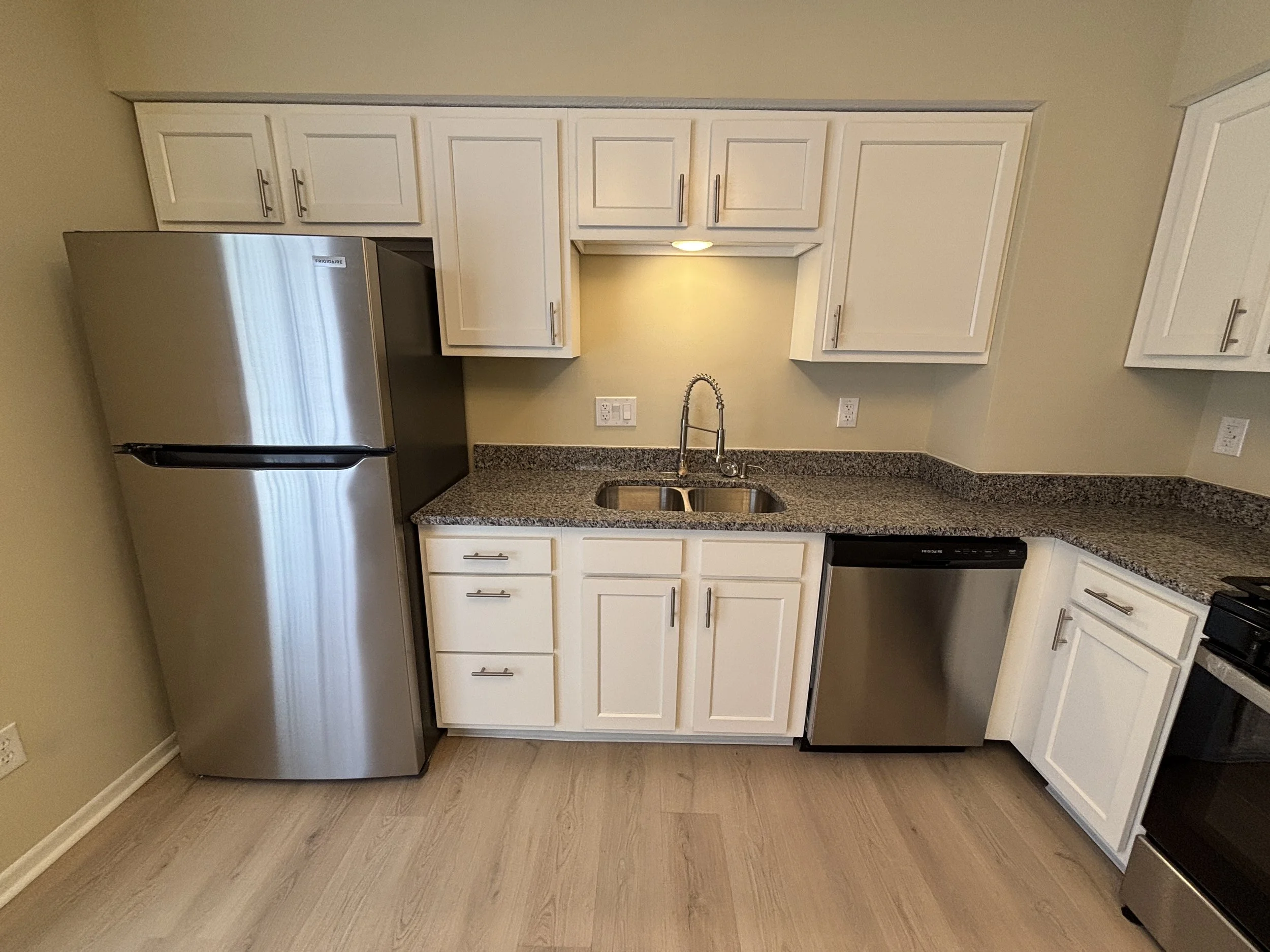 Kitchen with white cabinets, granite countertops, stainless steel refrigerator, dishwasher, and oven, beige walls, light wood flooring, and a double sink under a small cabinet with a light.