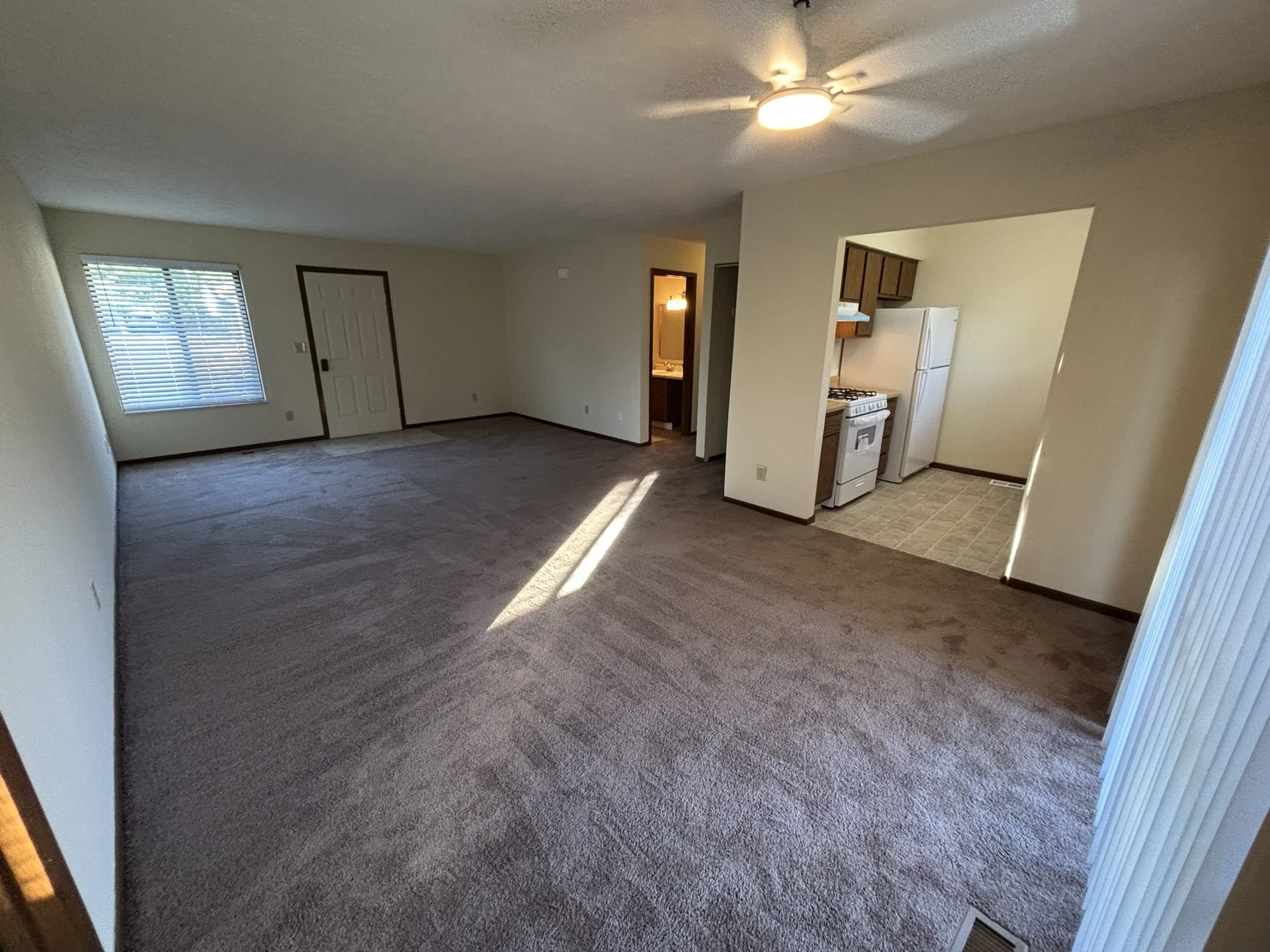 Empty living room with beige carpet, window with blinds, and an open view to a small kitchen with white appliances and brown cabinets.